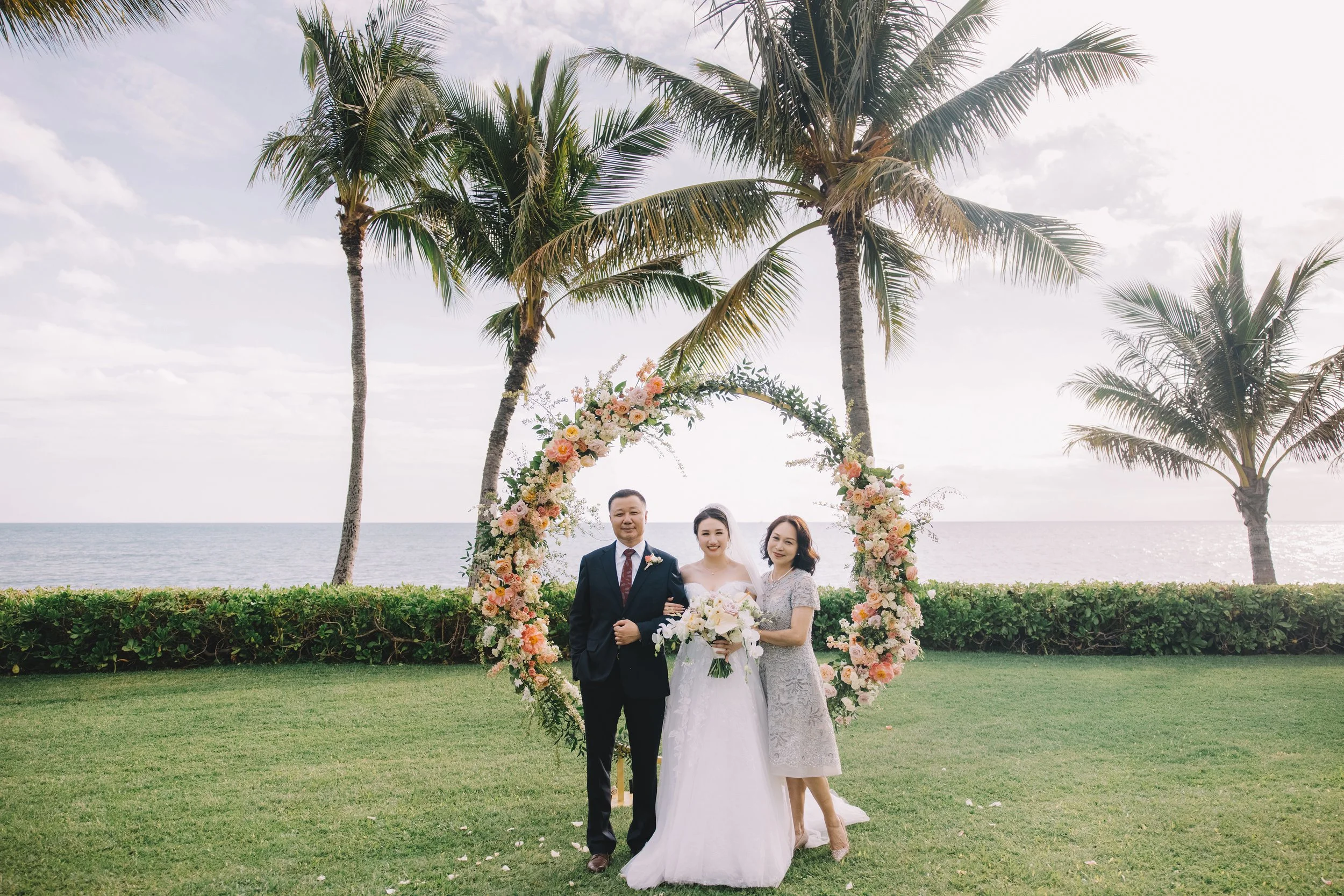 A wedding couple with a man and a woman beside the bride, standing outdoors on a grassy area near the ocean, with palm trees and a floral arch in the background.
