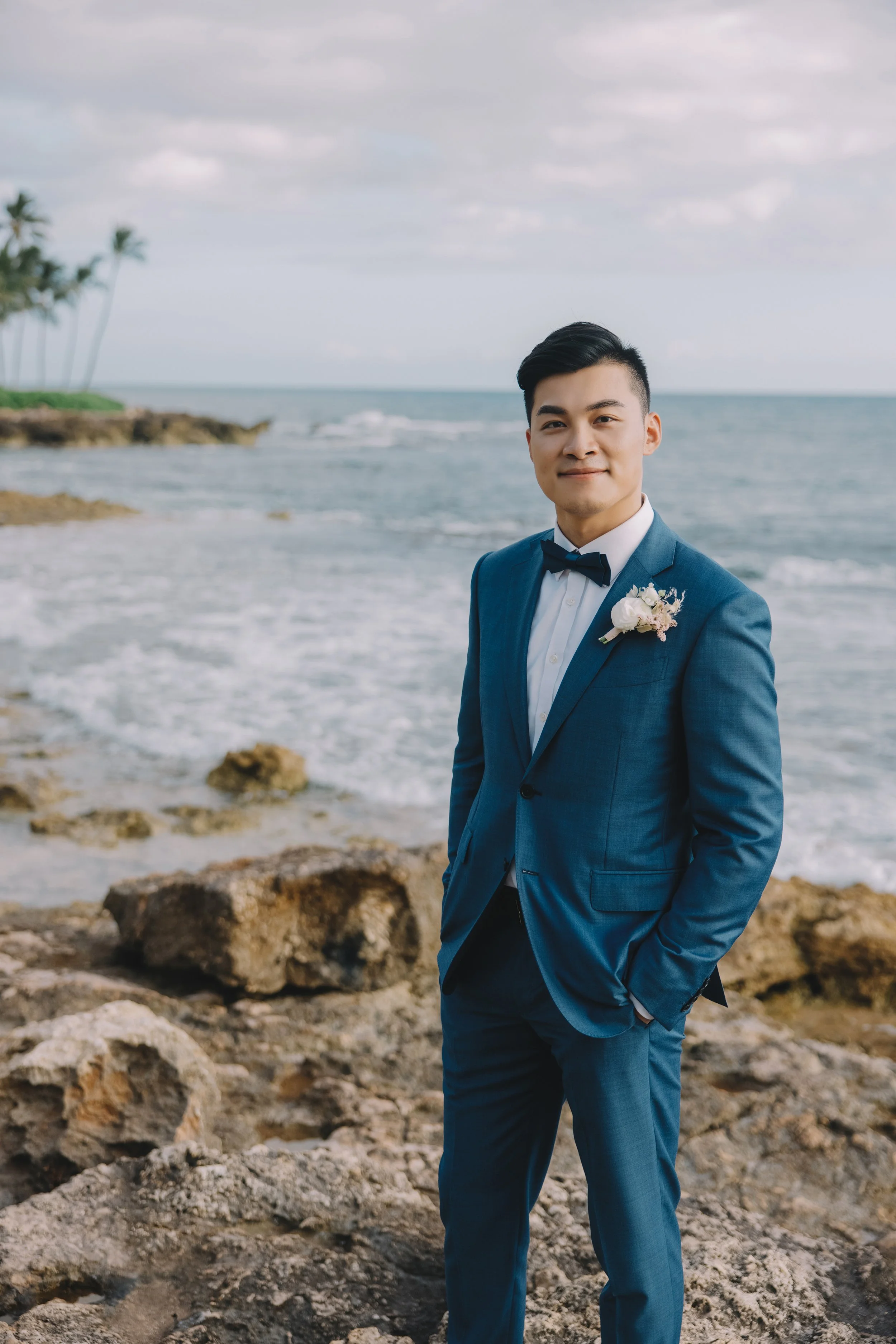 Young man in a blue suit with a flower boutonniere, standing near the rocky shoreline at a beach with ocean waves and palm trees in the background.
