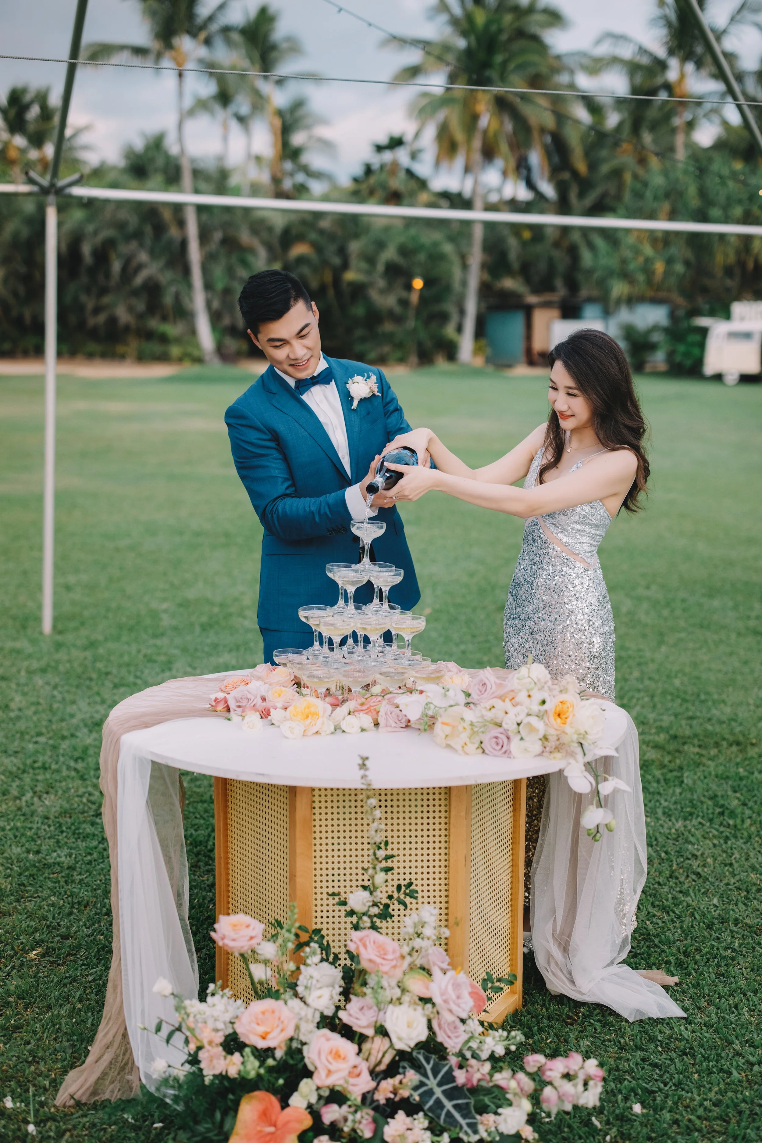 A couple in formal attire celebrating their wedding outdoors by pouring champagne into a pyramid of glasses on a decorated table with flowers, surrounded by palm trees.