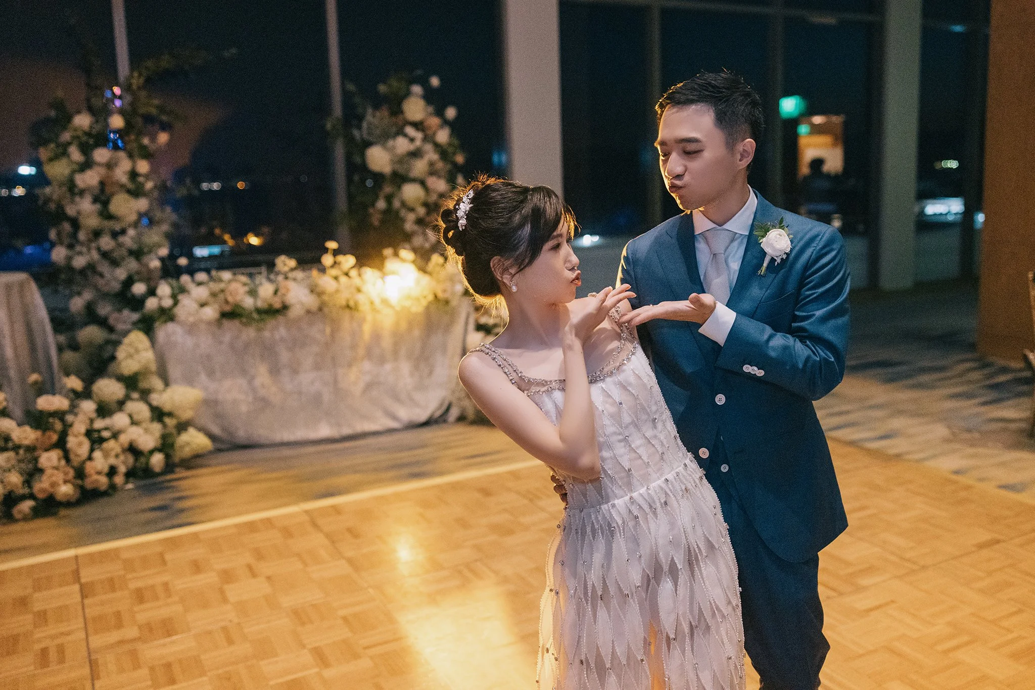 A bride and groom dancing at their wedding reception, with the bride in a light pink dress and the groom in a blue suit, on a wooden dance floor with floral decorations in the background.