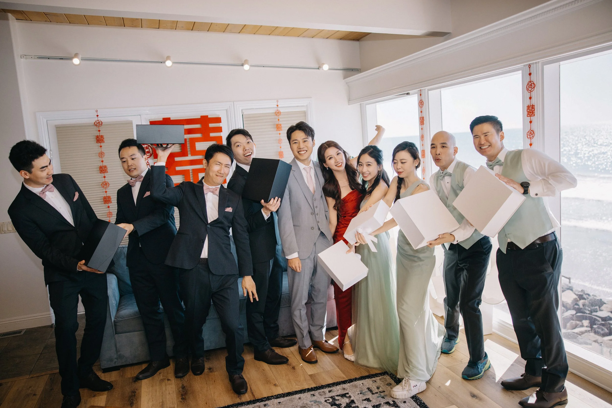 A group of people dressed in formal and semi-formal attire celebrating together indoors, holding gift boxes and smiling, with a sea view through large windows in the background.