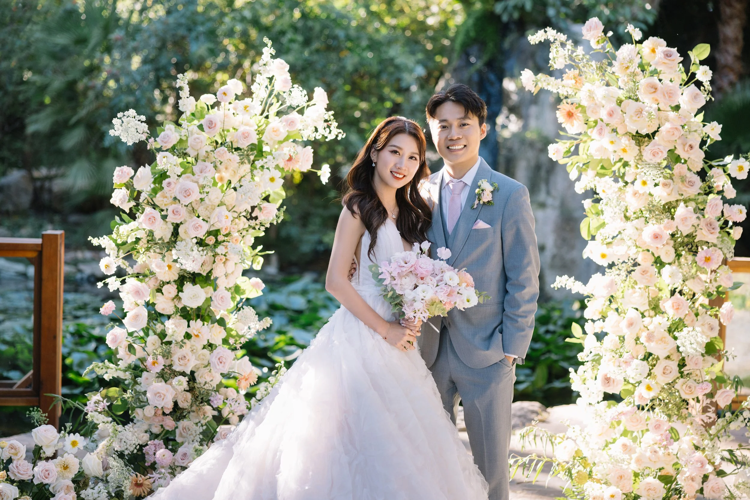 A bride and groom pose together outdoors at their wedding, surrounded by large floral arrangements of pink and white flowers, with green trees in the background.