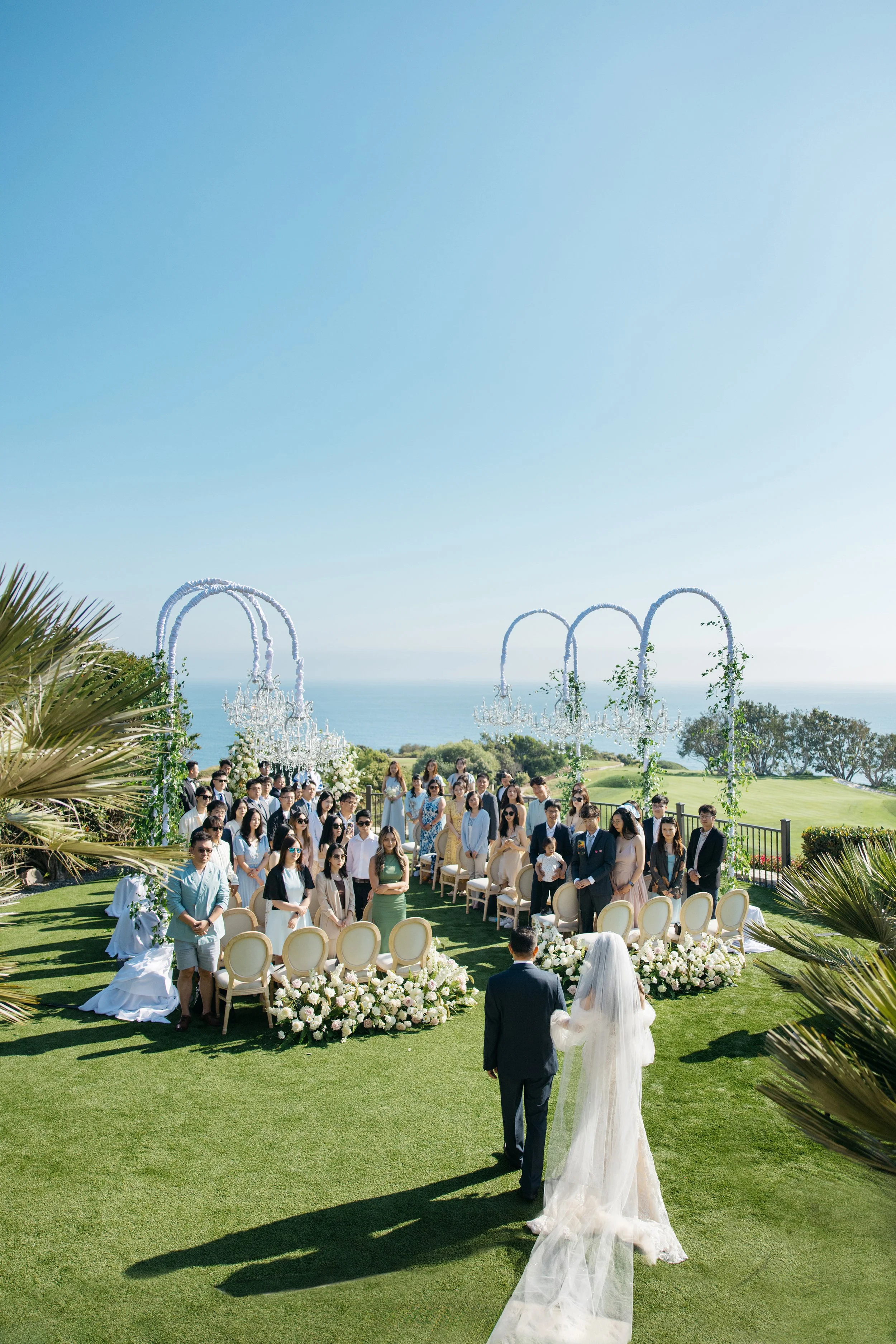 A wedding ceremony outdoors on a grassy lawn with an ocean view. Bride and groom walk hand in hand towards seated guests, with guests standing behind decorated chairs and floral arrangements. Overhead, white arches with hanging chandeliers and greene