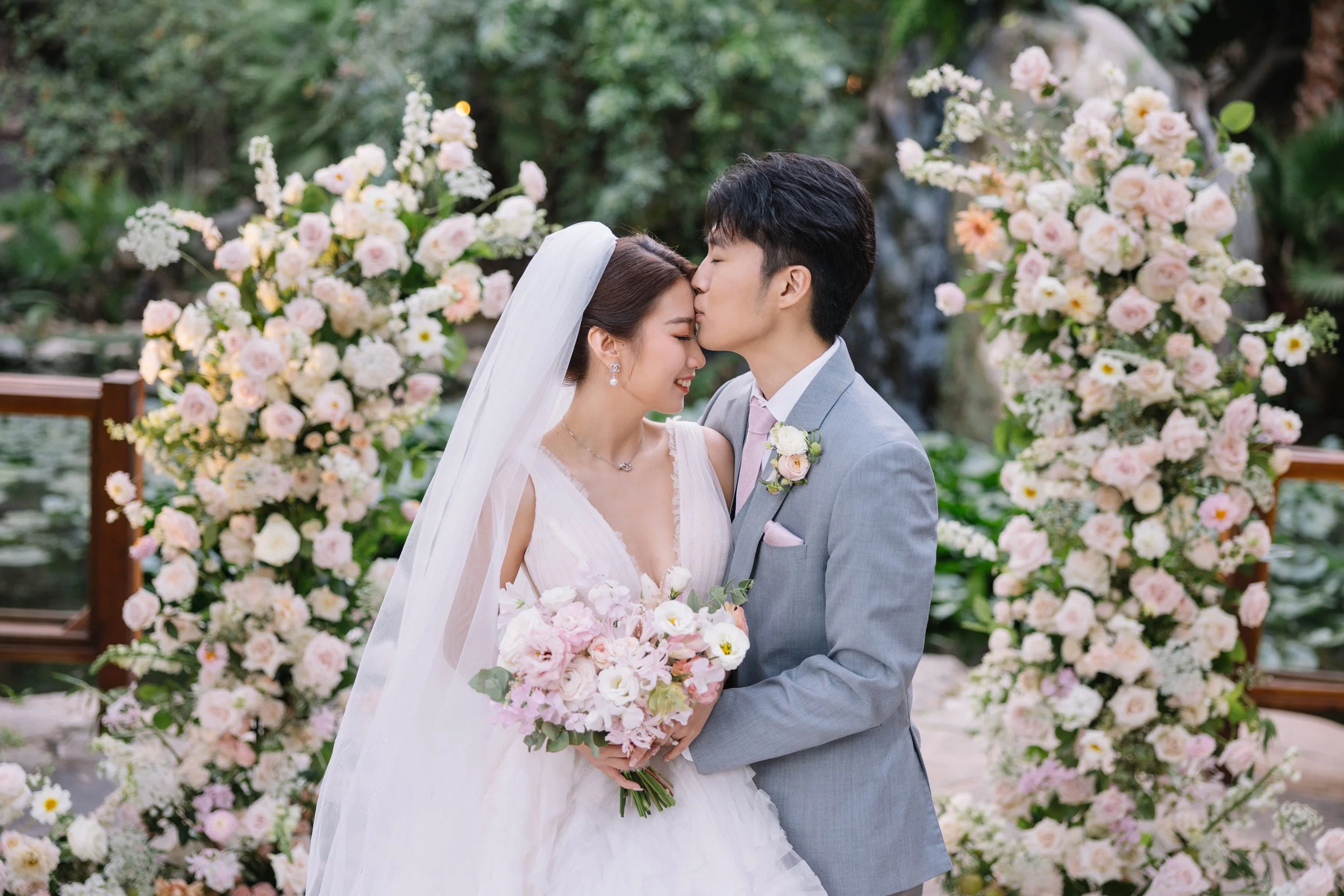 A bride and groom share a kiss at their wedding, surrounded by large floral arrangements of pink and white roses and other flowers in a lush outdoor setting.