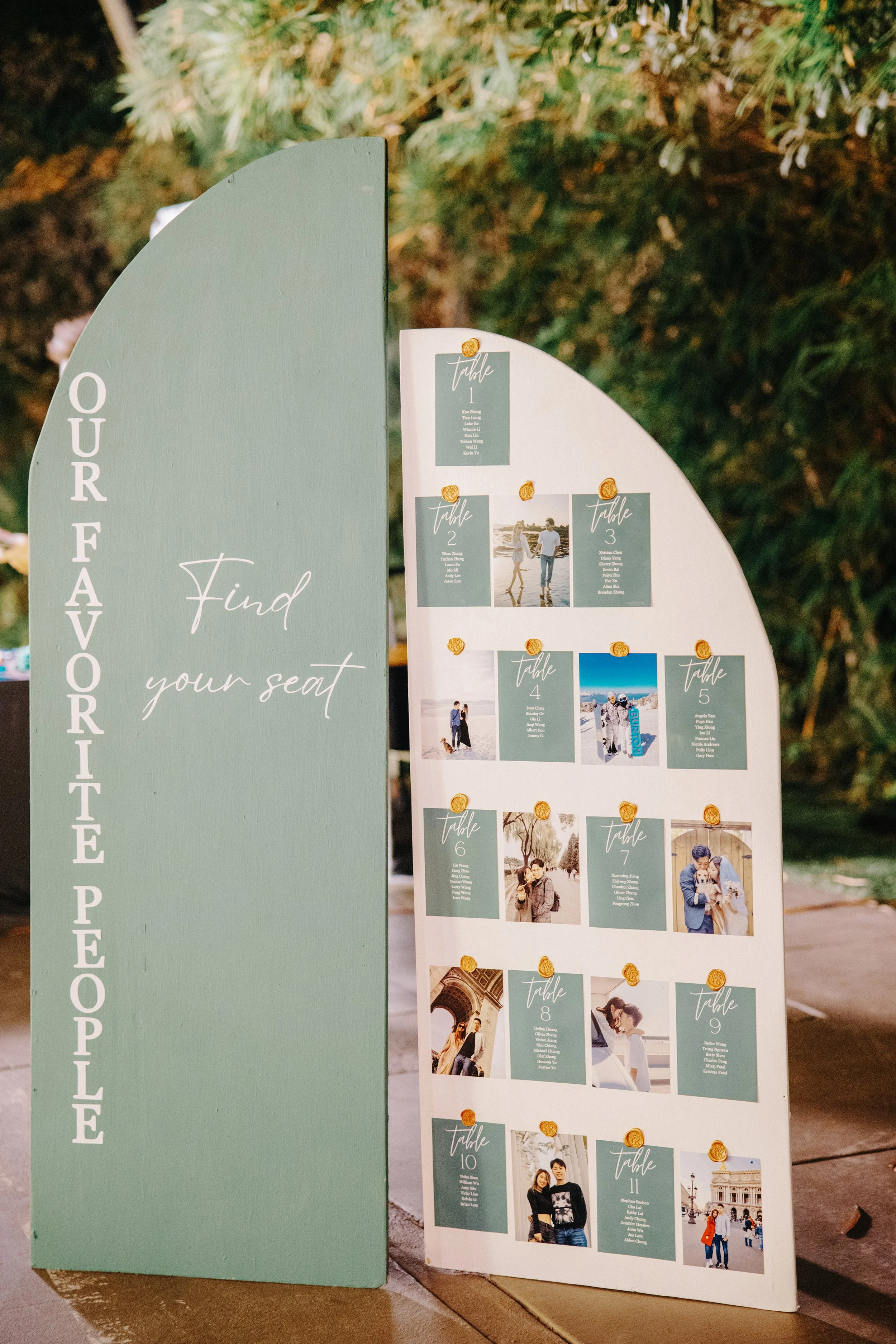 A green and white seating chart board for a wedding, with table numbers and guest names, set outdoors surrounded by greenery.