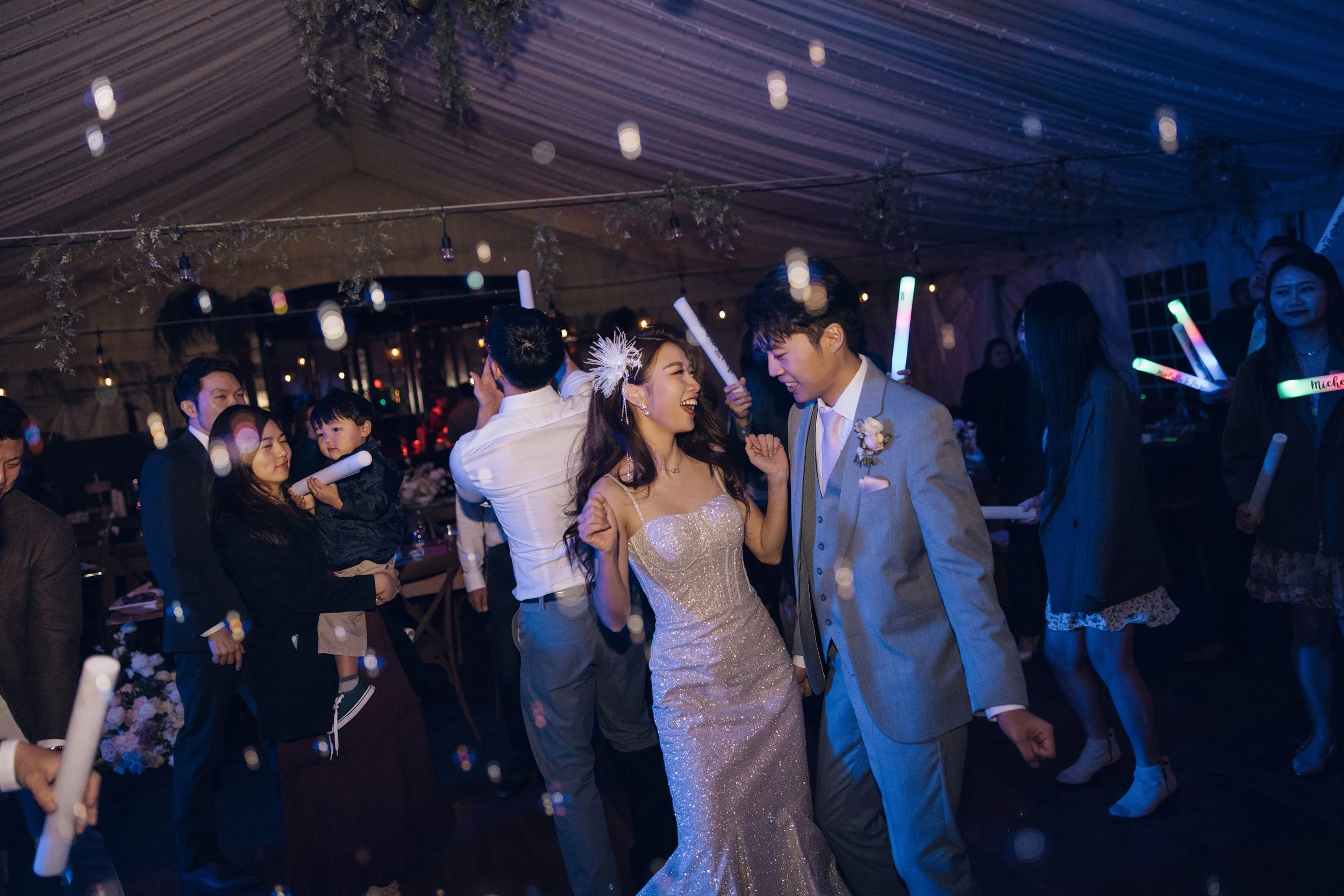 Couple dancing at wedding reception, surrounded by friends and family, under a decorated tent with string lights.