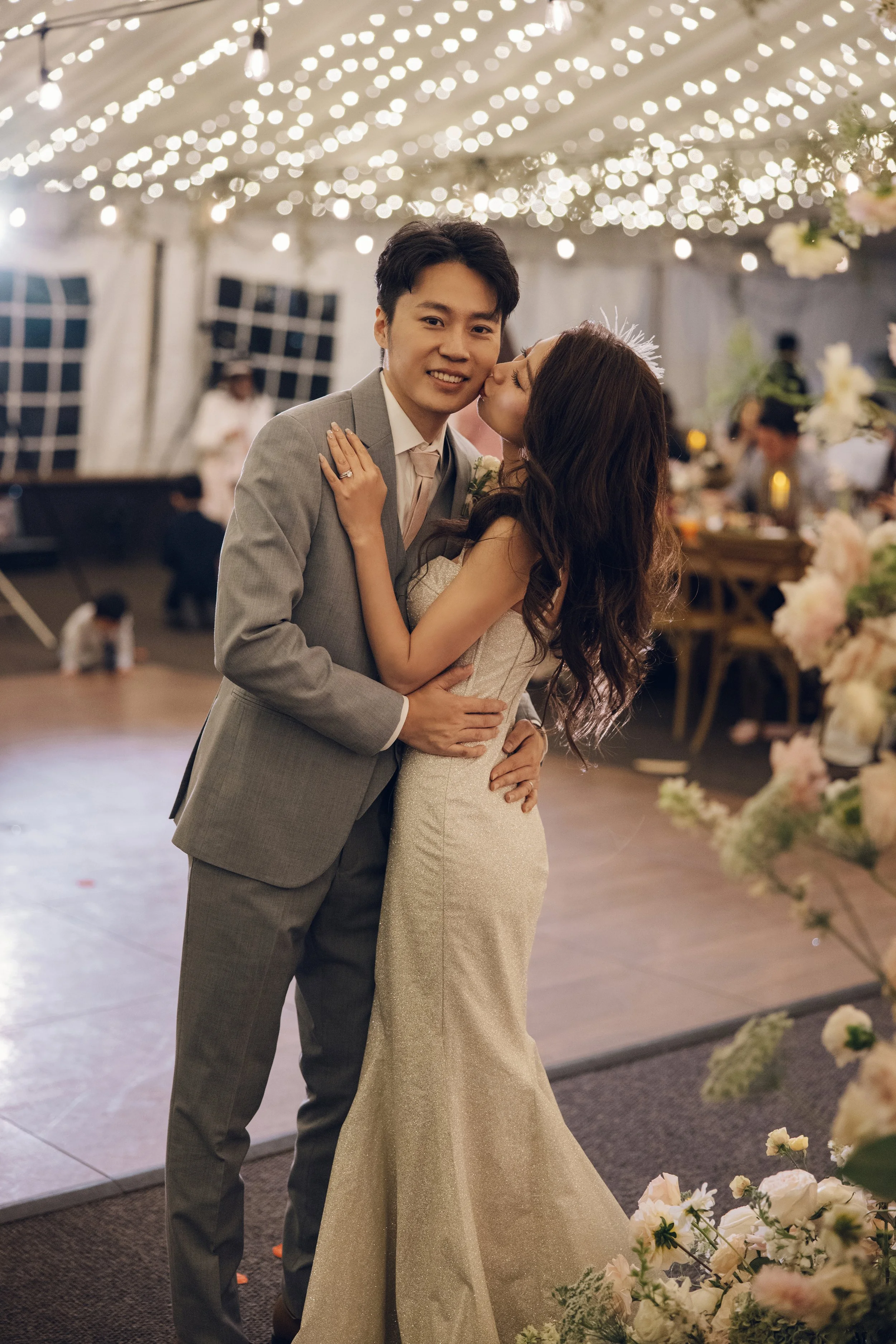A newlywed couple sharing a kiss at their wedding reception, decorated with string lights and flowers.