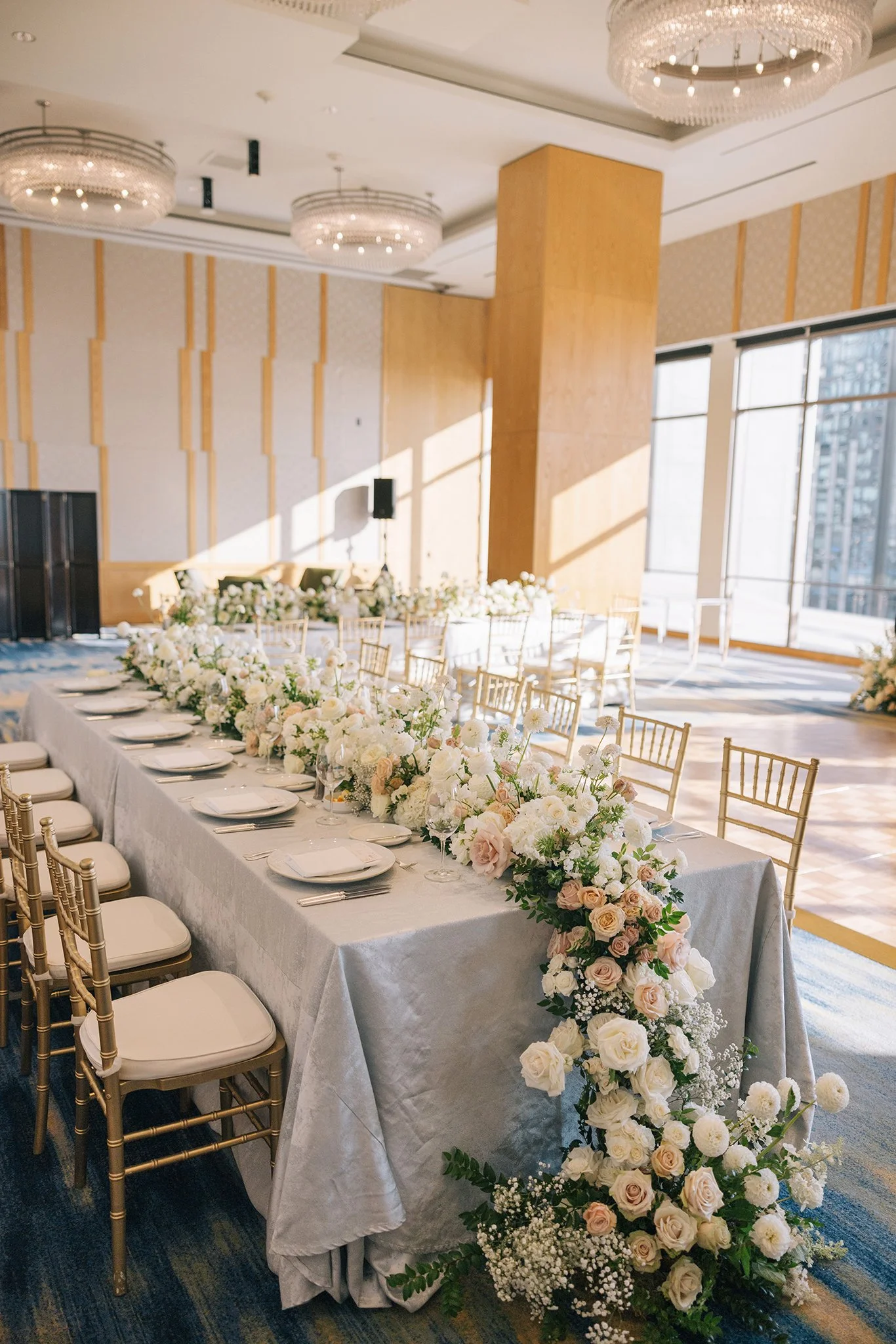 A banquet table decorated with a floral arrangement of white and blush roses, white ranunculus, baby's breath, and greenery, set for a wedding or event, with gold chairs and white plates in a bright, high-ceilinged room with large windows.