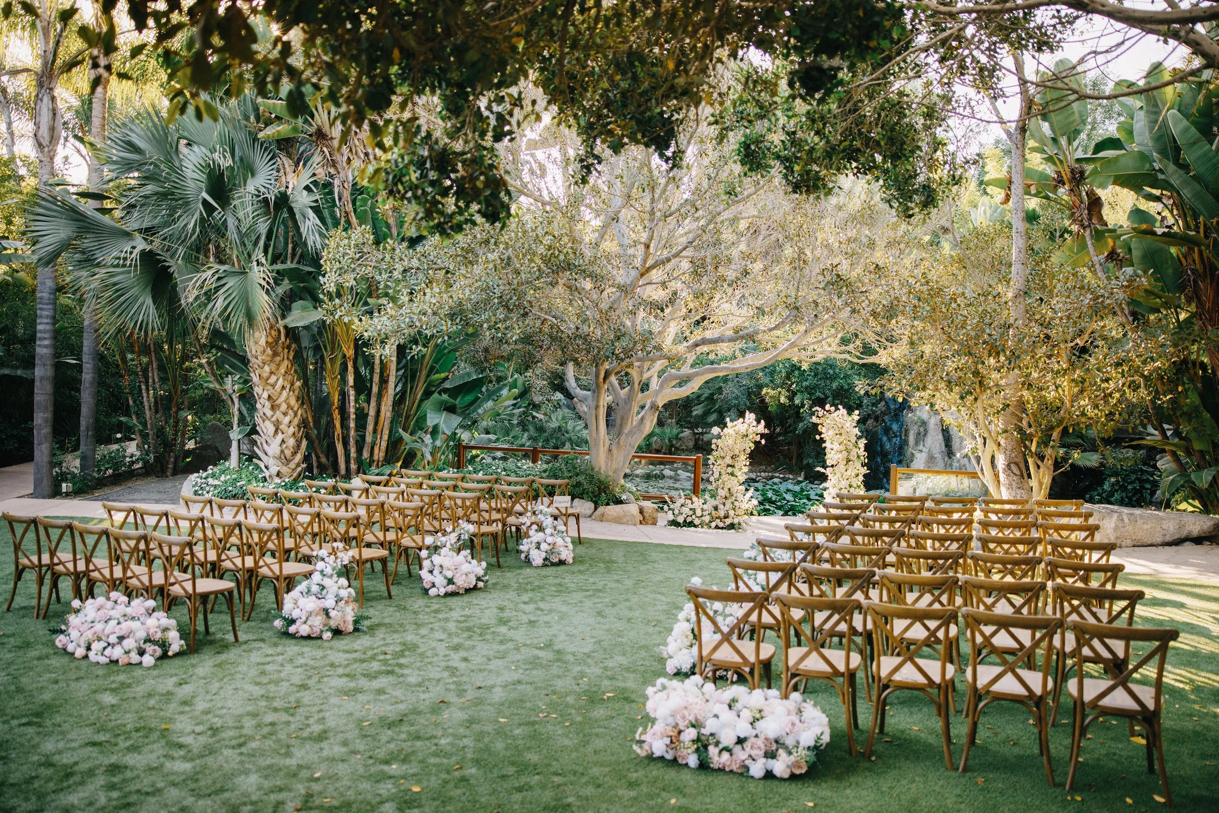 Outdoor wedding ceremony setup with rows of wooden chairs and floral arrangements on a grassy lawn surrounded by lush green trees.