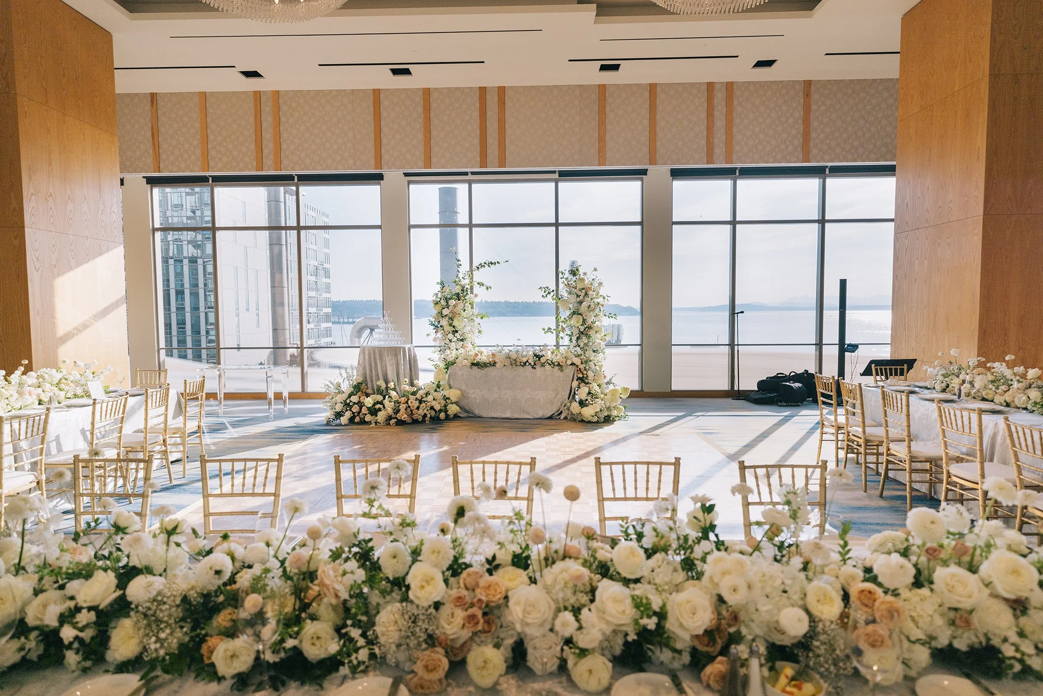 Wedding ceremony setup in a bright room with large windows overlooking a body of water. The room features a floral arch at the front, tables with floral arrangements, and gold chairs arranged for guests.