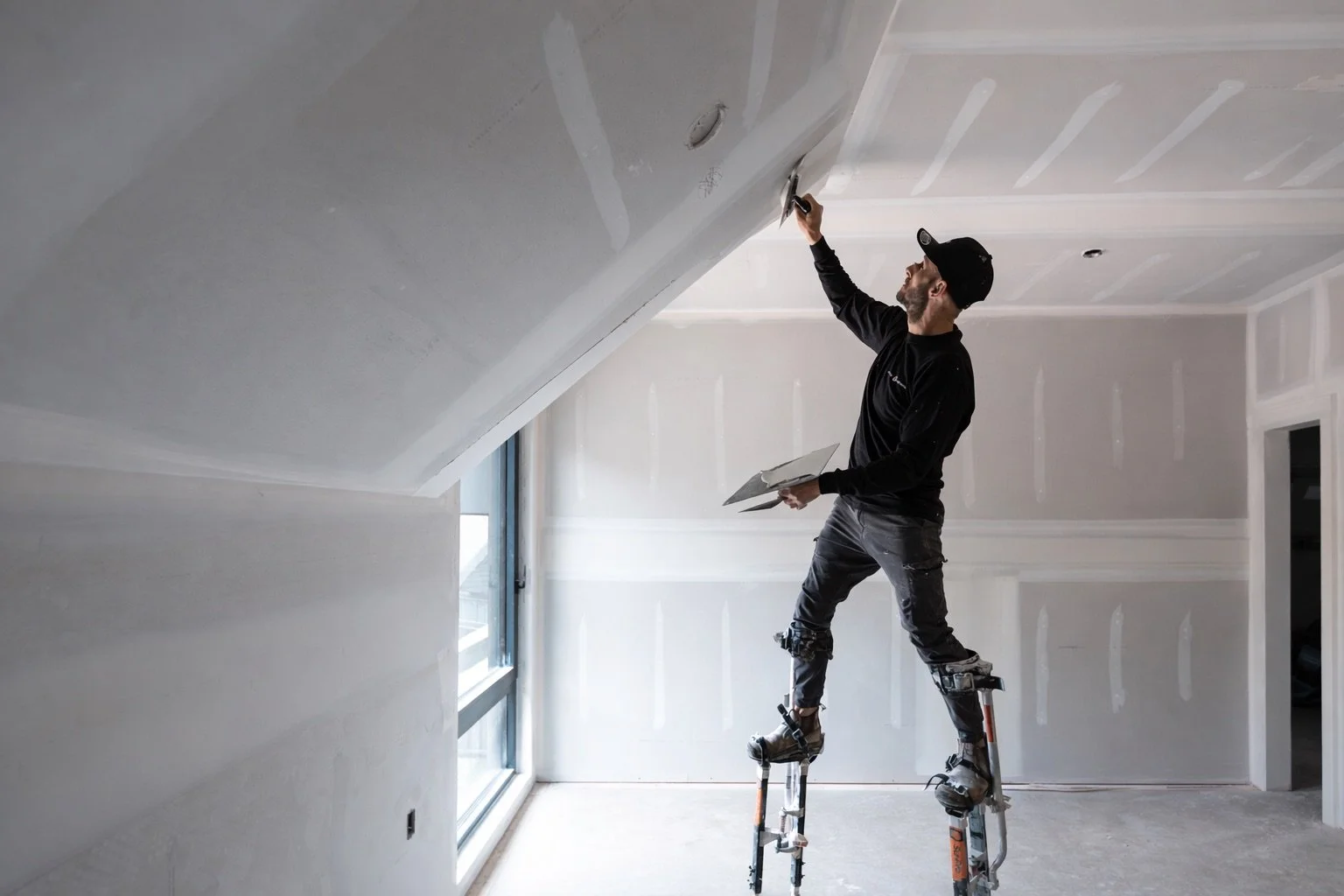 A man with a beard, wearing a black cap, black long-sleeve shirt, and gray pants working on drywall in an unfinished room with sloped ceiling. He is standing on stilts and sanding the drywall ceiling, holding a trowel in one hand and a drywall knife in the other.