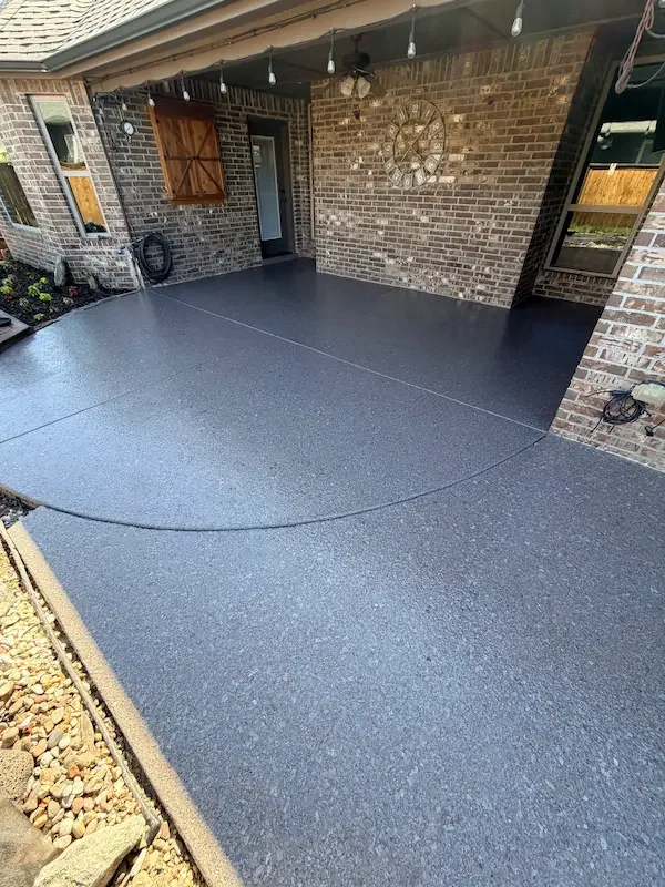 Freshly poured and sealed concrete patio with a textured finish on a brick house porch, decorated with string lights and a large clock on the wall.