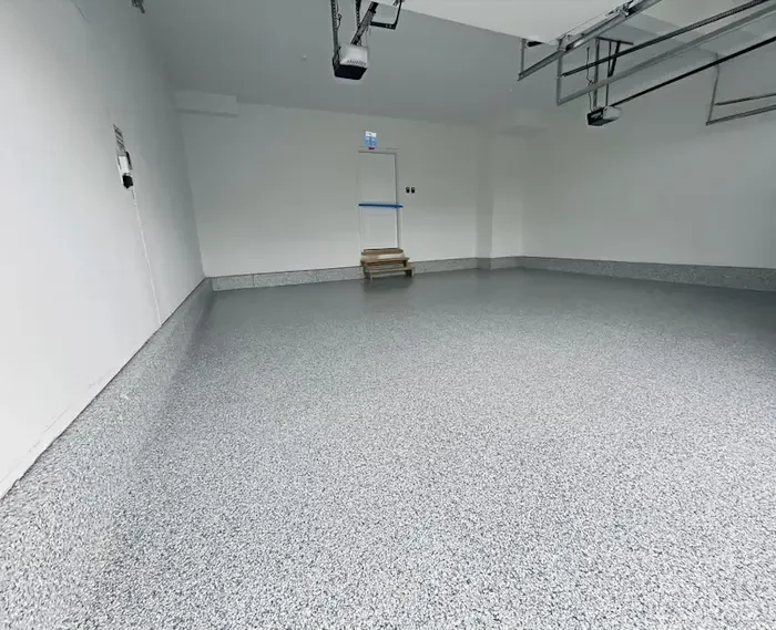 Empty garage with gray speckled epoxy floor, white walls, and a closed door with a step ladder in front.
