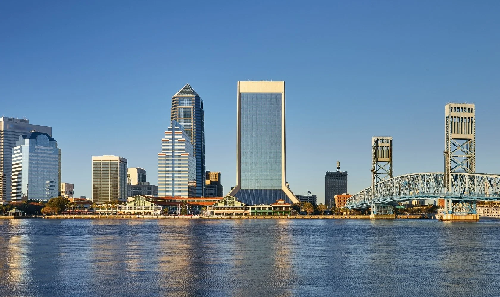 Skyline of downtown Jacksonville, Florida, with modern skyscrapers, a bridge on the right, and water in the foreground.