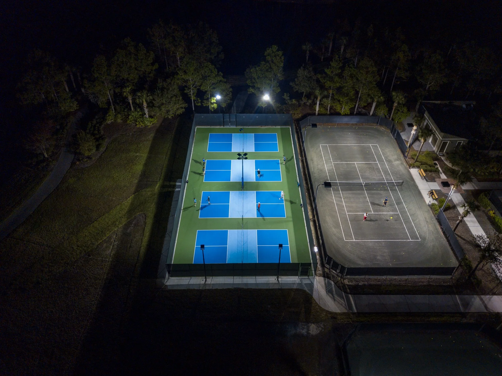 An aerial nighttime view of a tennis court with players, surrounded by a fence, located near a building and trees, with bright lights illuminating the court.