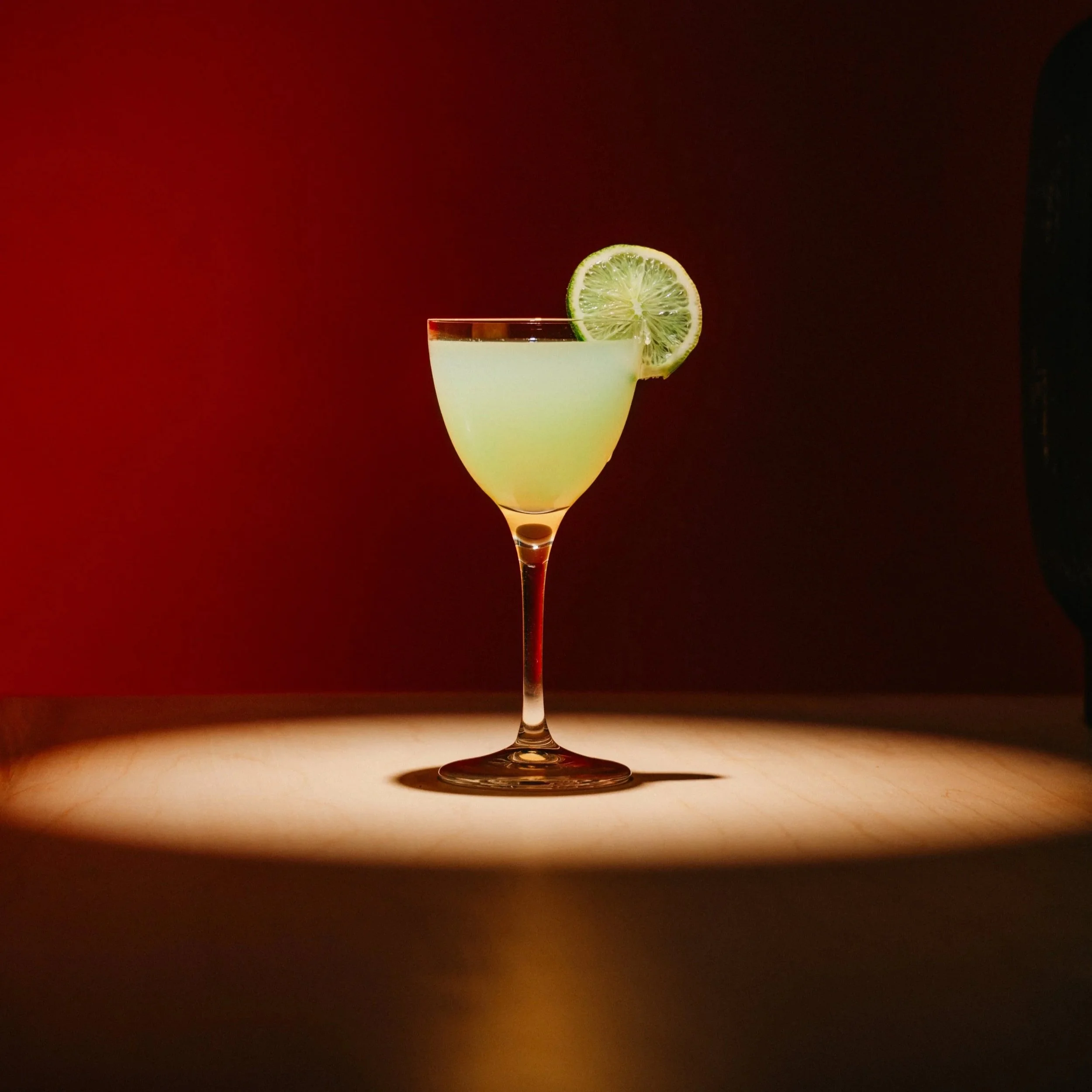 A cocktail glass with a pale yellow drink garnished with a slice of lime, set against a dark red background with a spotlight on the glass.