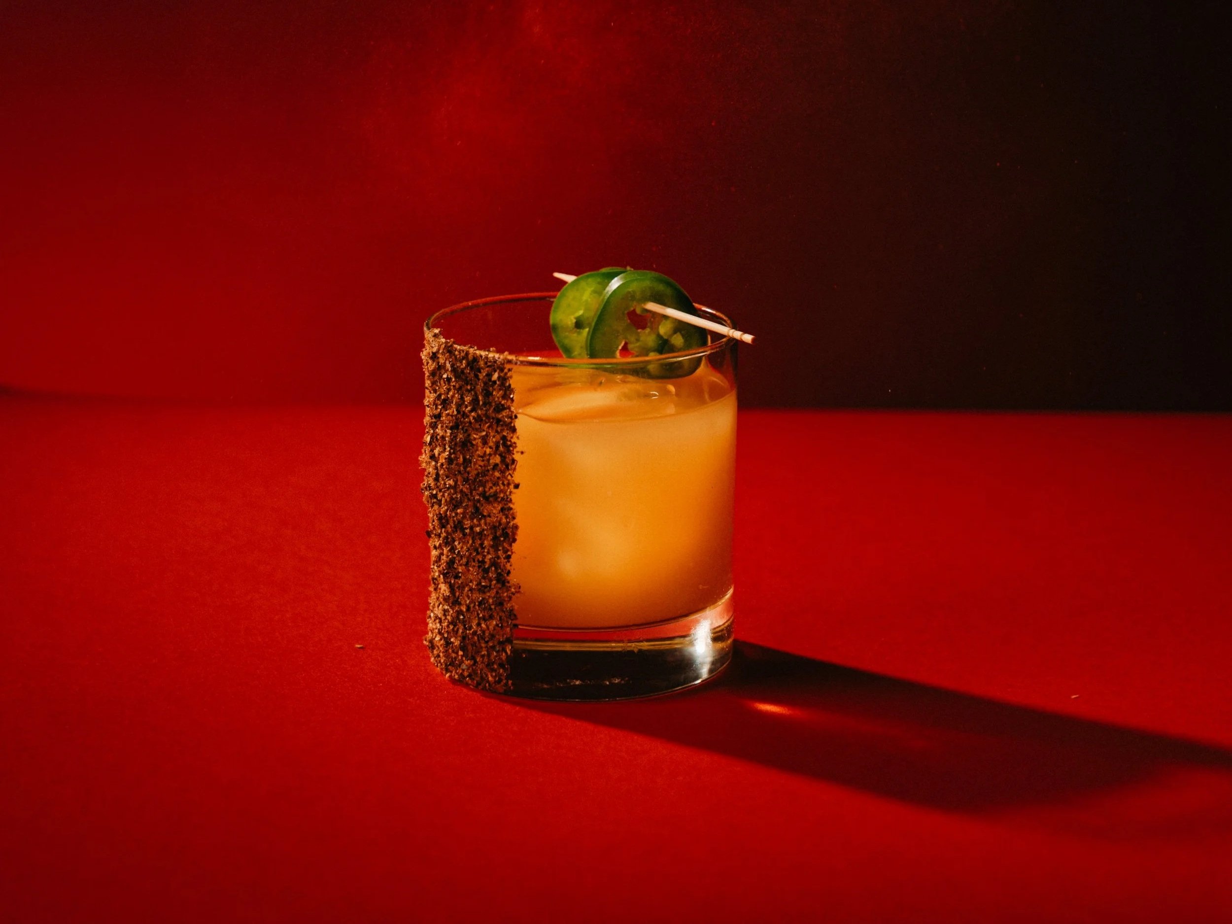 A cocktail glass with a yellow drink, garnished with a jalapeño pepper and a rim coated with chili powder, against a dark and red background.