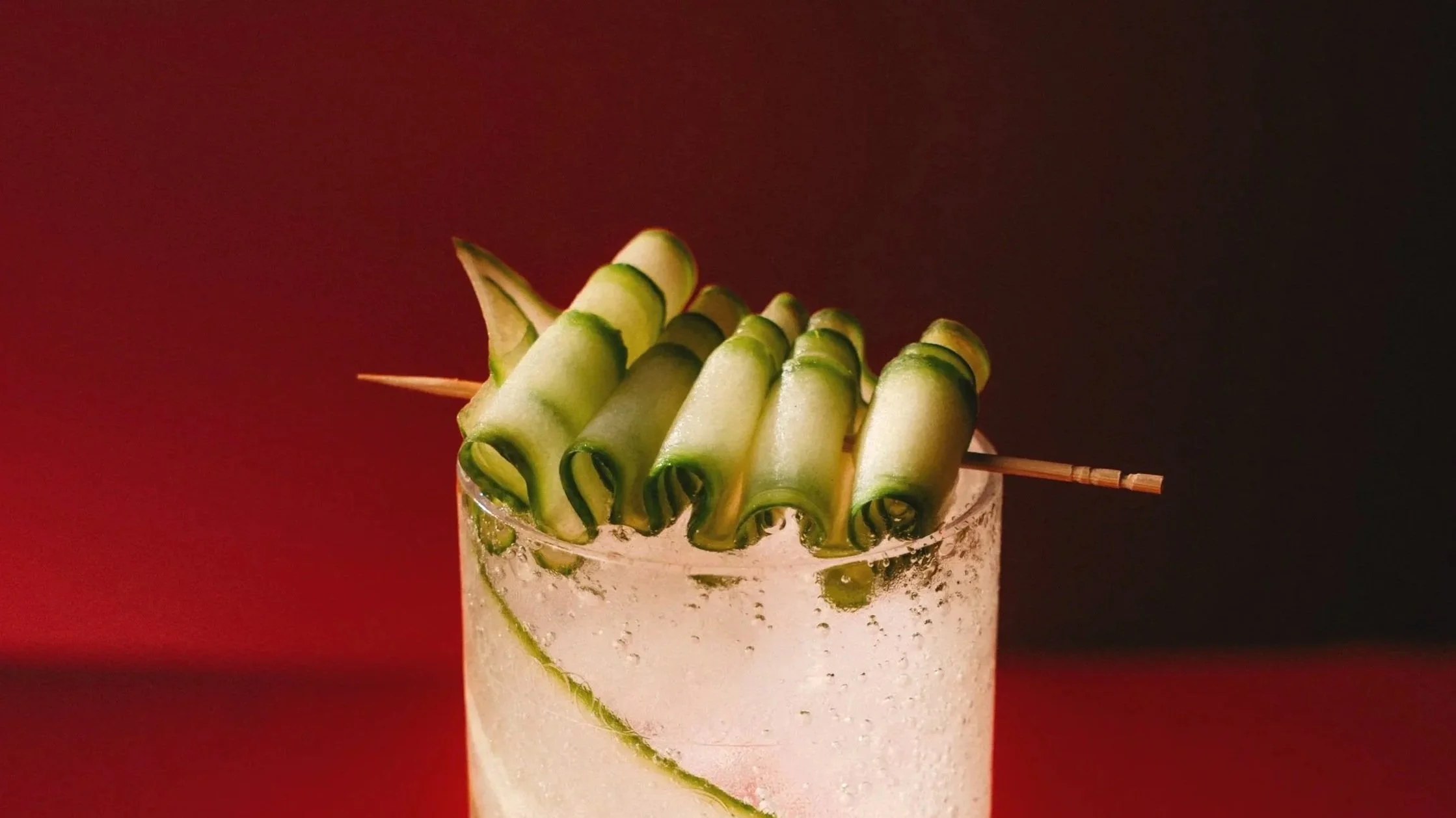 A cocktail glass with a clear liquid, garnished with a spiraled cucumber and a cucumber spear, against a red and black gradient background.