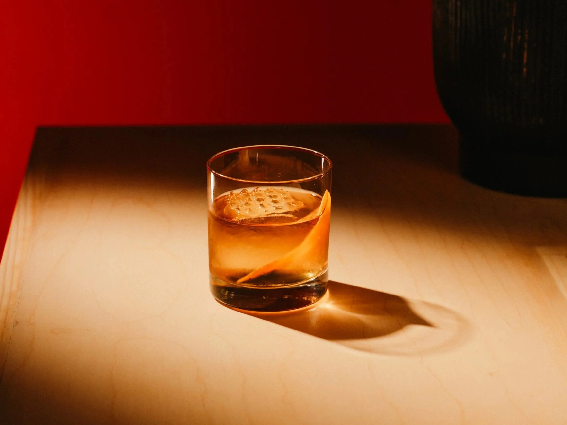 A glass of whiskey with ice and an orange peel on a wooden table, casting a shadow.