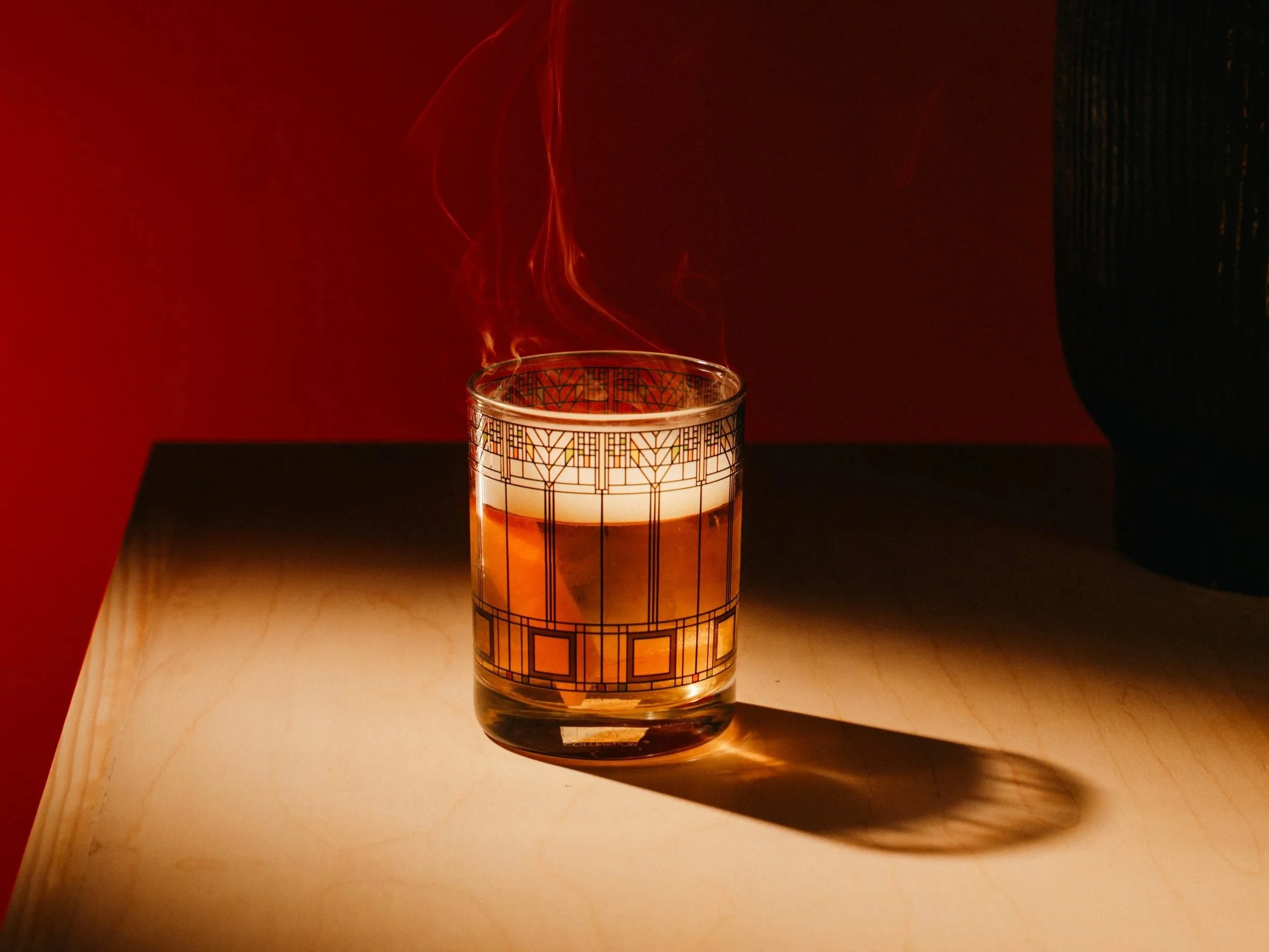 A lit scented candle with a decorative geometric pattern on a glass container, casting a shadow on a wooden surface, against a red background.