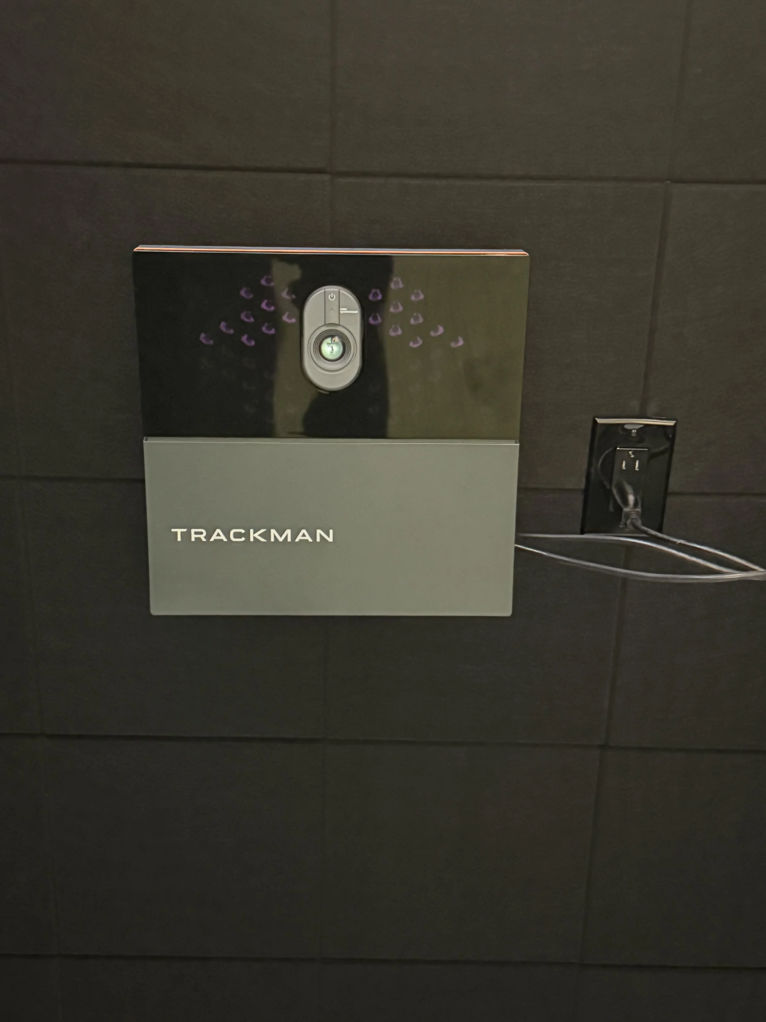A black and gray wall-mounted device labeled 'Trackman' with a small camera or sensor on top, connected to an outlet on a dark tiled wall.