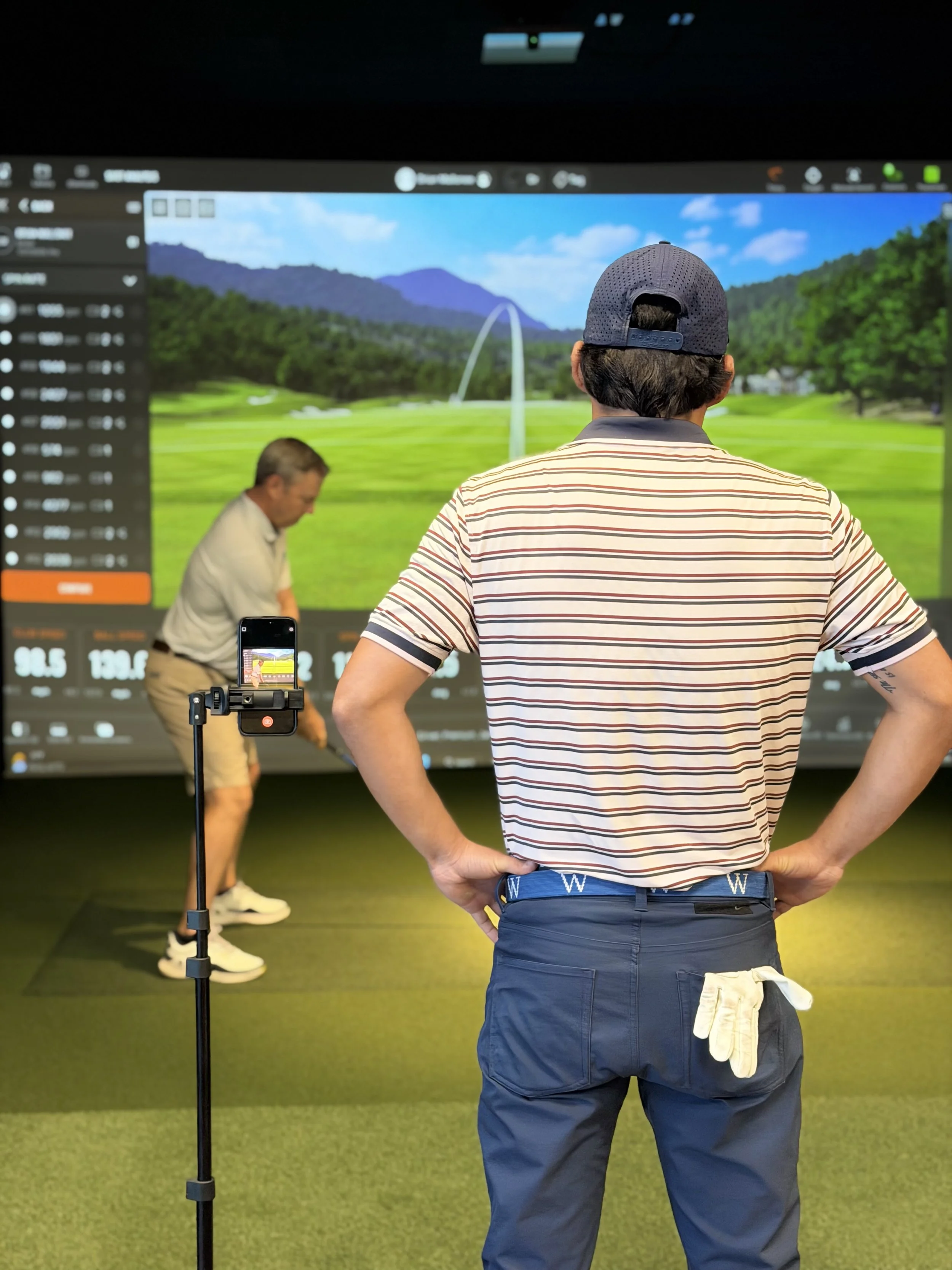 A man stands with his back facing the camera, observing a golf simulation on a large screen, with a golf course image and a man preparing to swing in front of the screen, and a smartphone mounted on a tripod recording the scene.