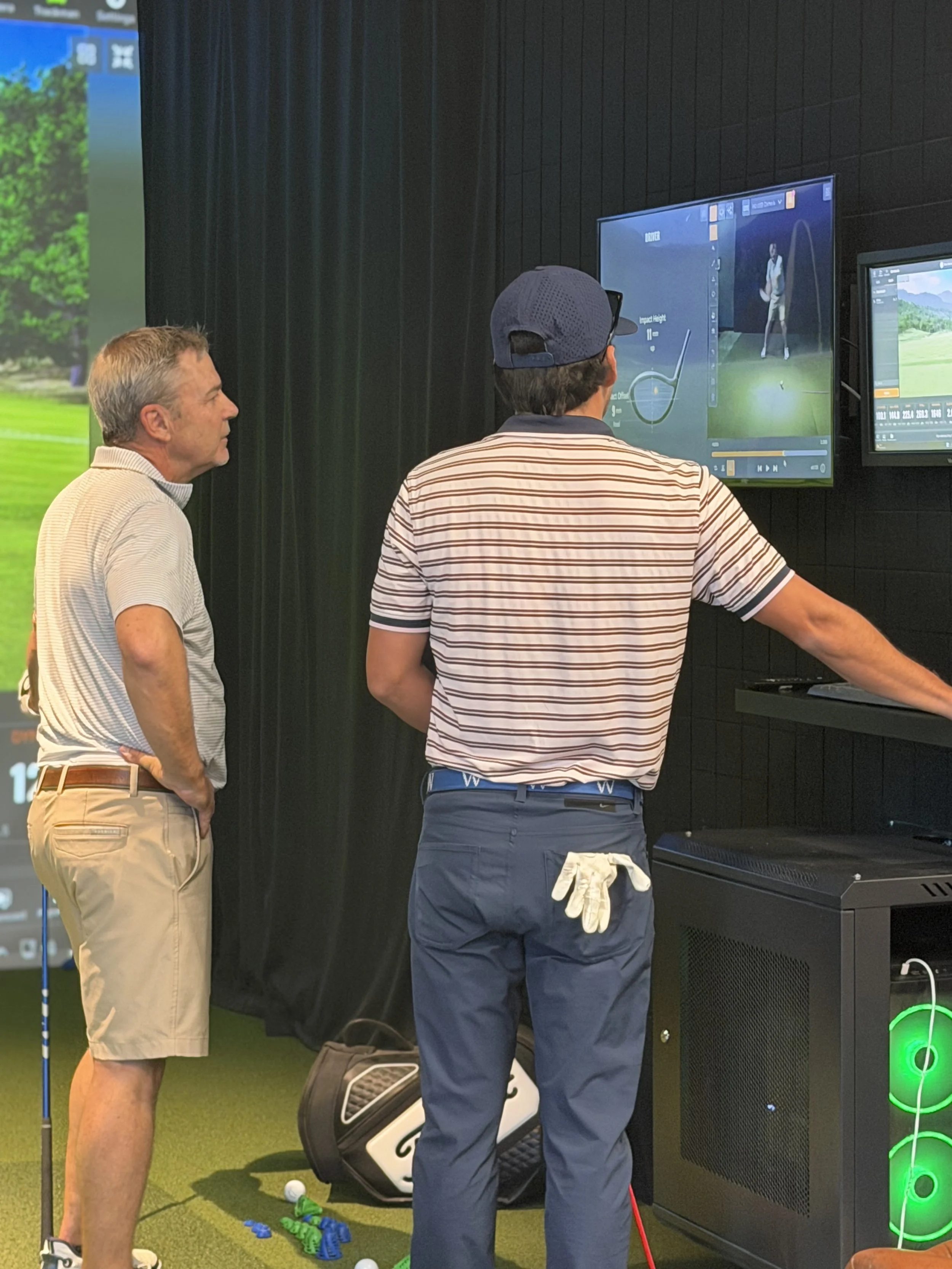 Two men are watching a golf simulator on a screen, one with a golf club in hand and a golf bag on the floor nearby.