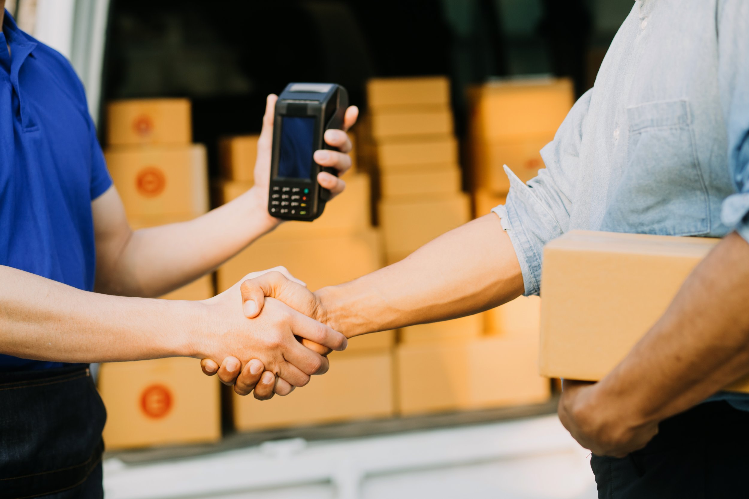 Two people shaking hands in front of shelves of cardboard boxes, one person holding a point-of-sale terminal.