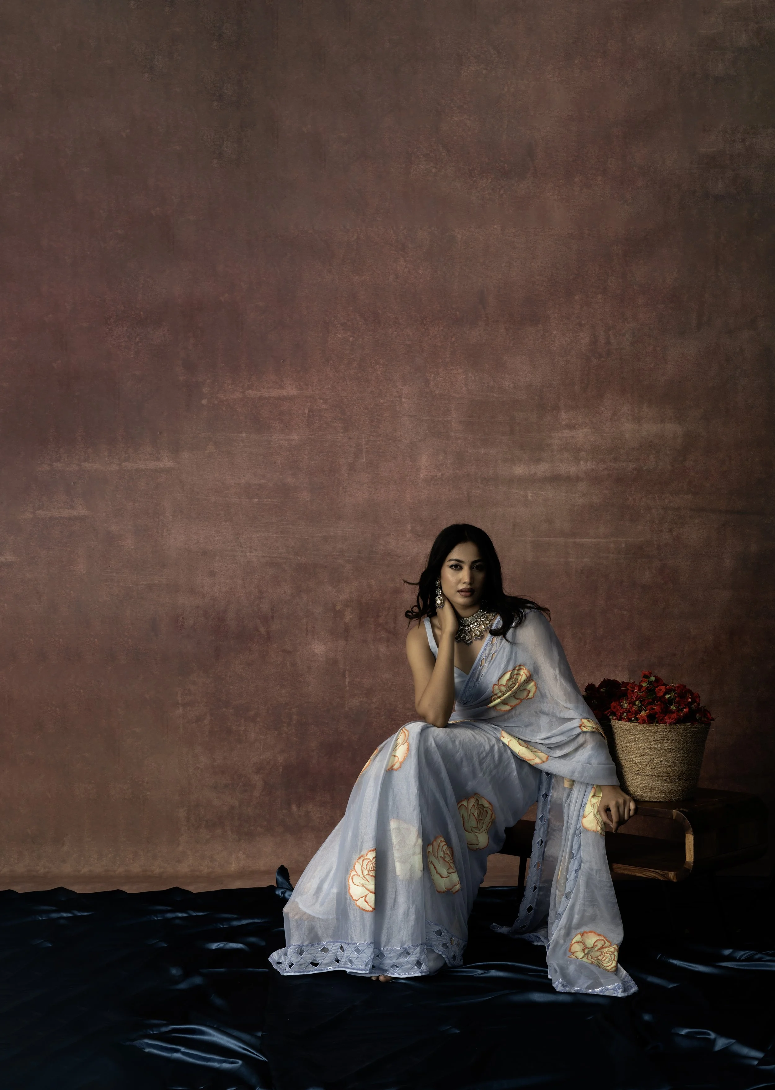 A woman in a traditional sari with embroidered flower patterns, sitting on the floor, next to a table with a basket of red roses, against a textured brown background.