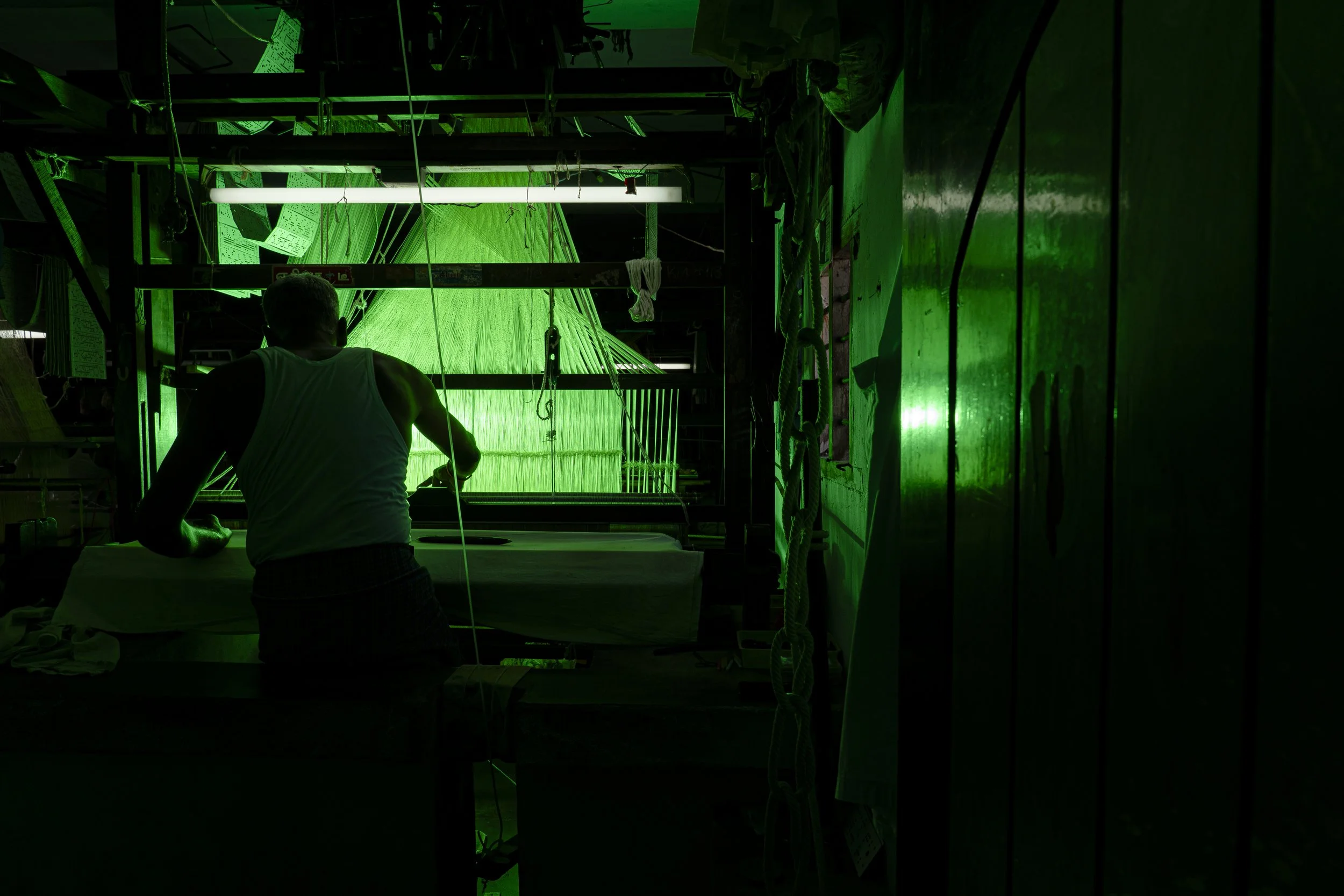 A person working at a fabric or textile workshop illuminated by green LED lighting. The silhouette of the person is visible, and they are handling fabric or material on a table.