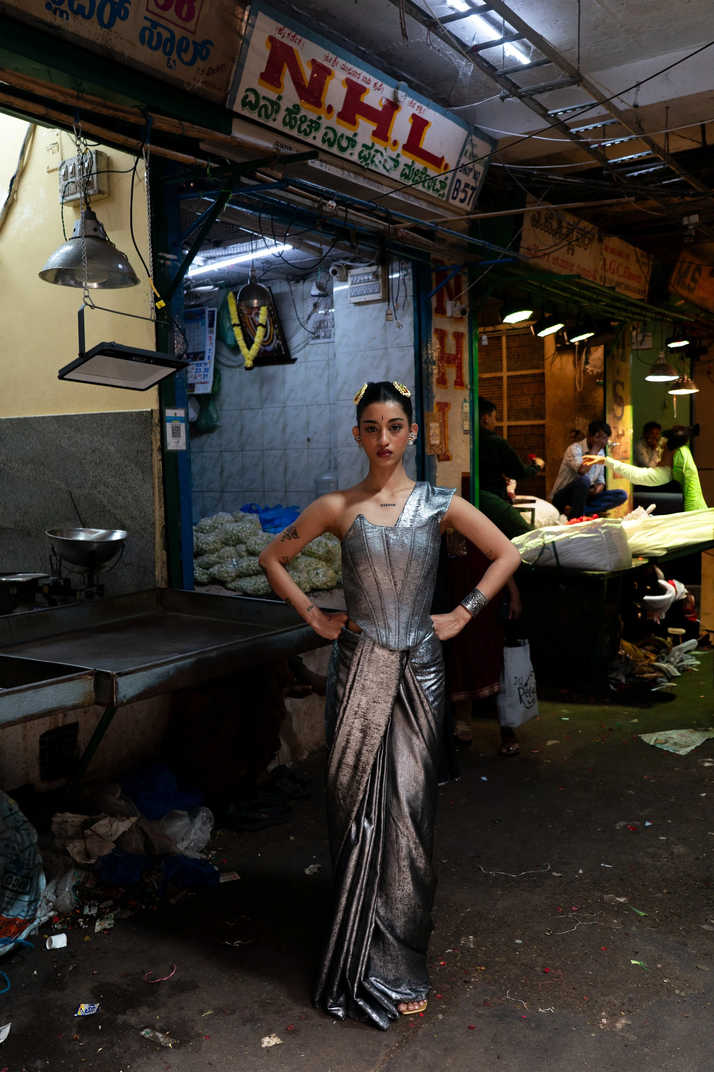 A woman wearing a shiny silver saree and jewelry stands confidently with her hands on her hips in a busy marketplace, with vendors and shoppers in the background.