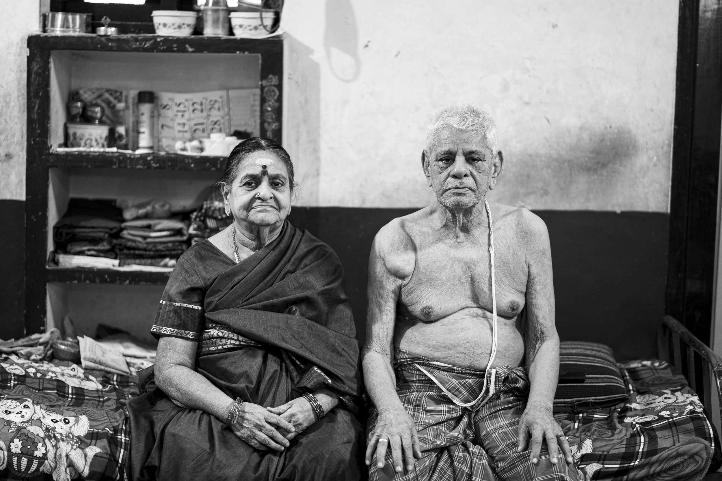 An elderly woman and elderly man sitting on a bed in a modest room. The woman is dressed in traditional clothing, and the man is shirtless wearing a lungi. Both have serious expressions.