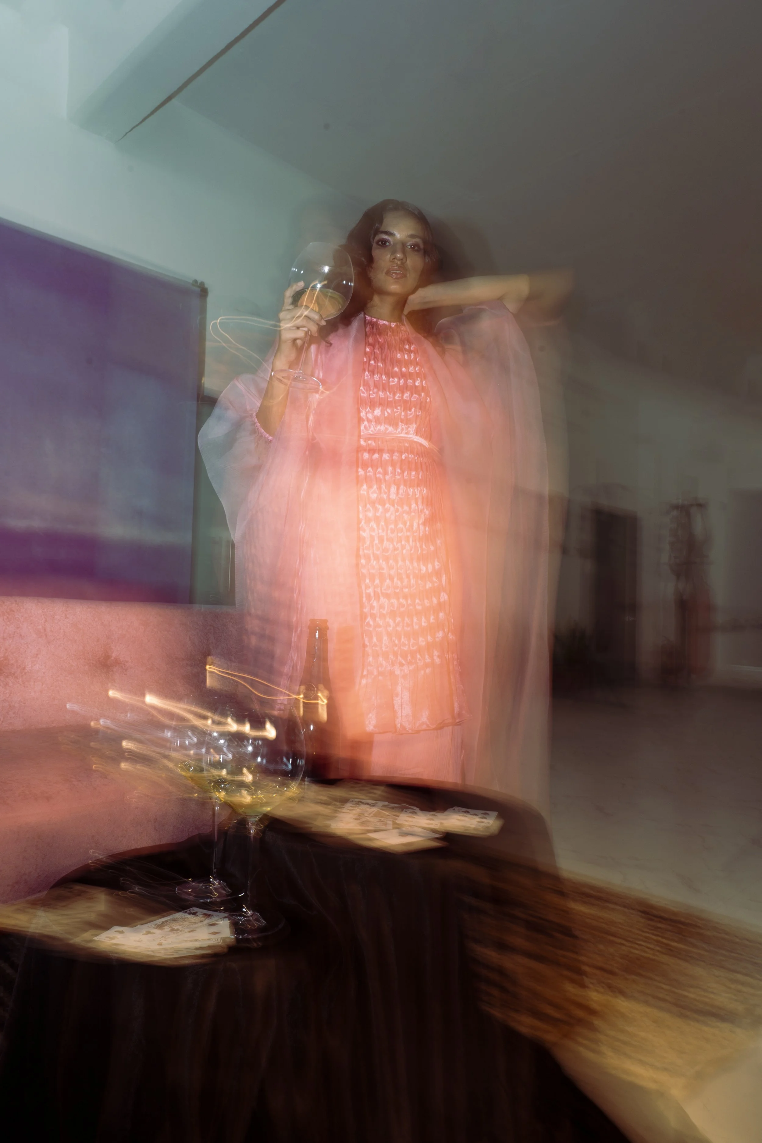 A woman wearing a pink dress and matching pink jacket standing indoors, holding a wine glass in her right hand, with a blurred effect due to motion.