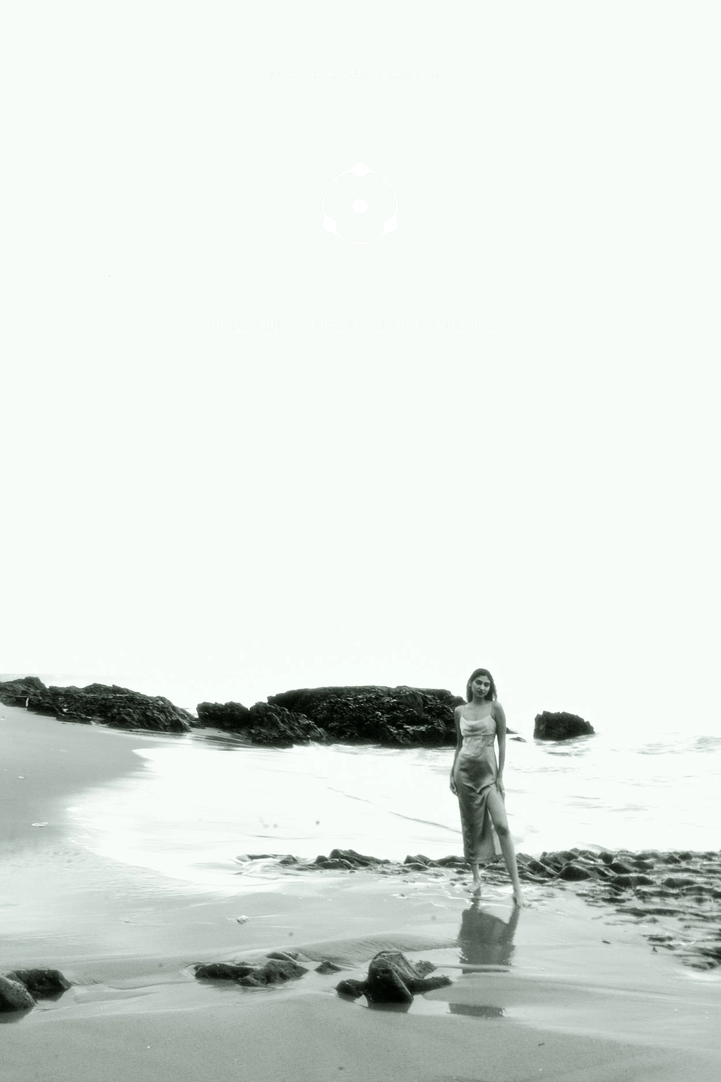 Black and white photo of a woman standing on a sandy beach near rocks, with the ocean in the background.