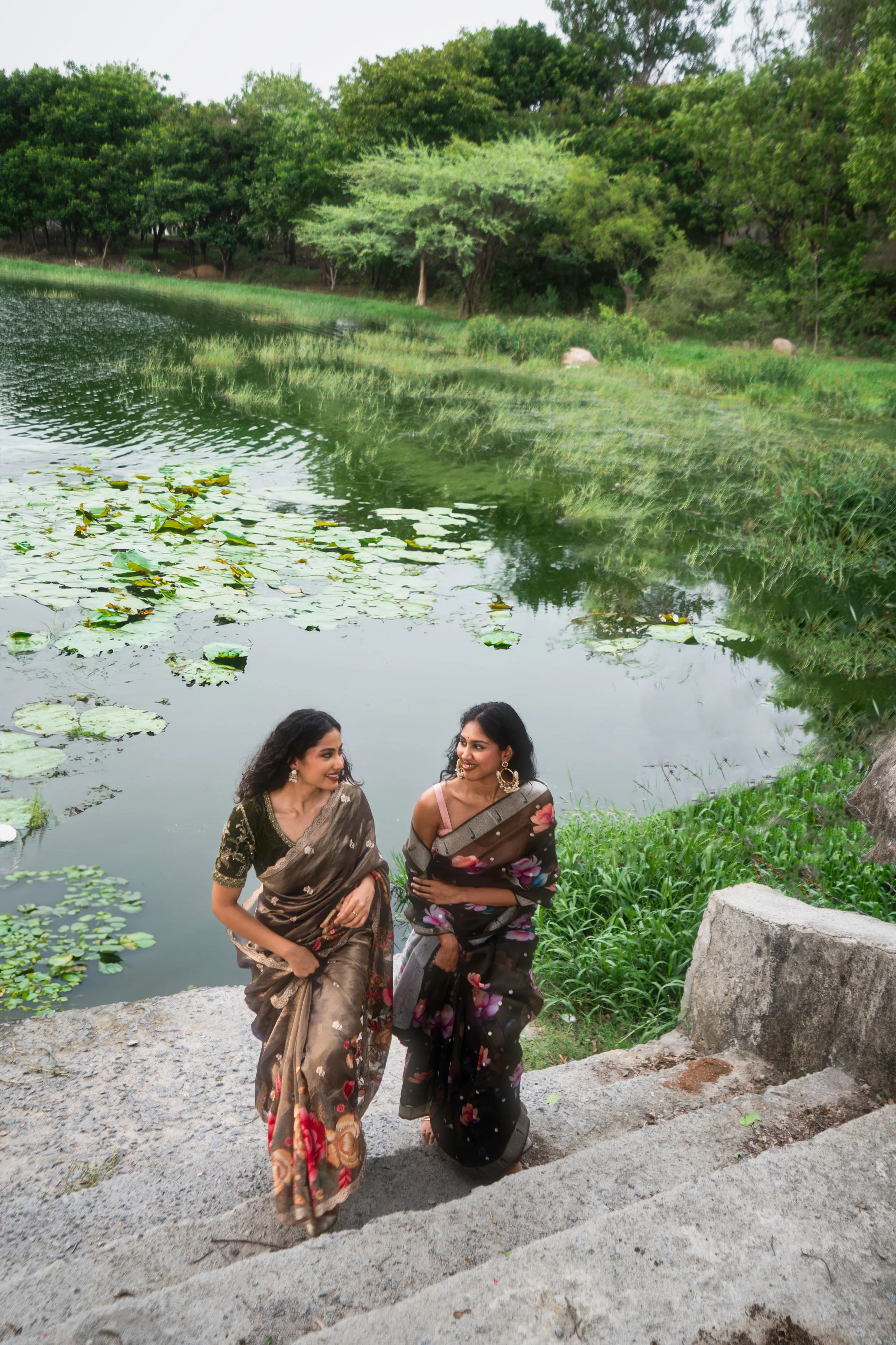Two women in sarees walking down stone stairs beside a pond with water lilies, surrounded by trees and greenery.
