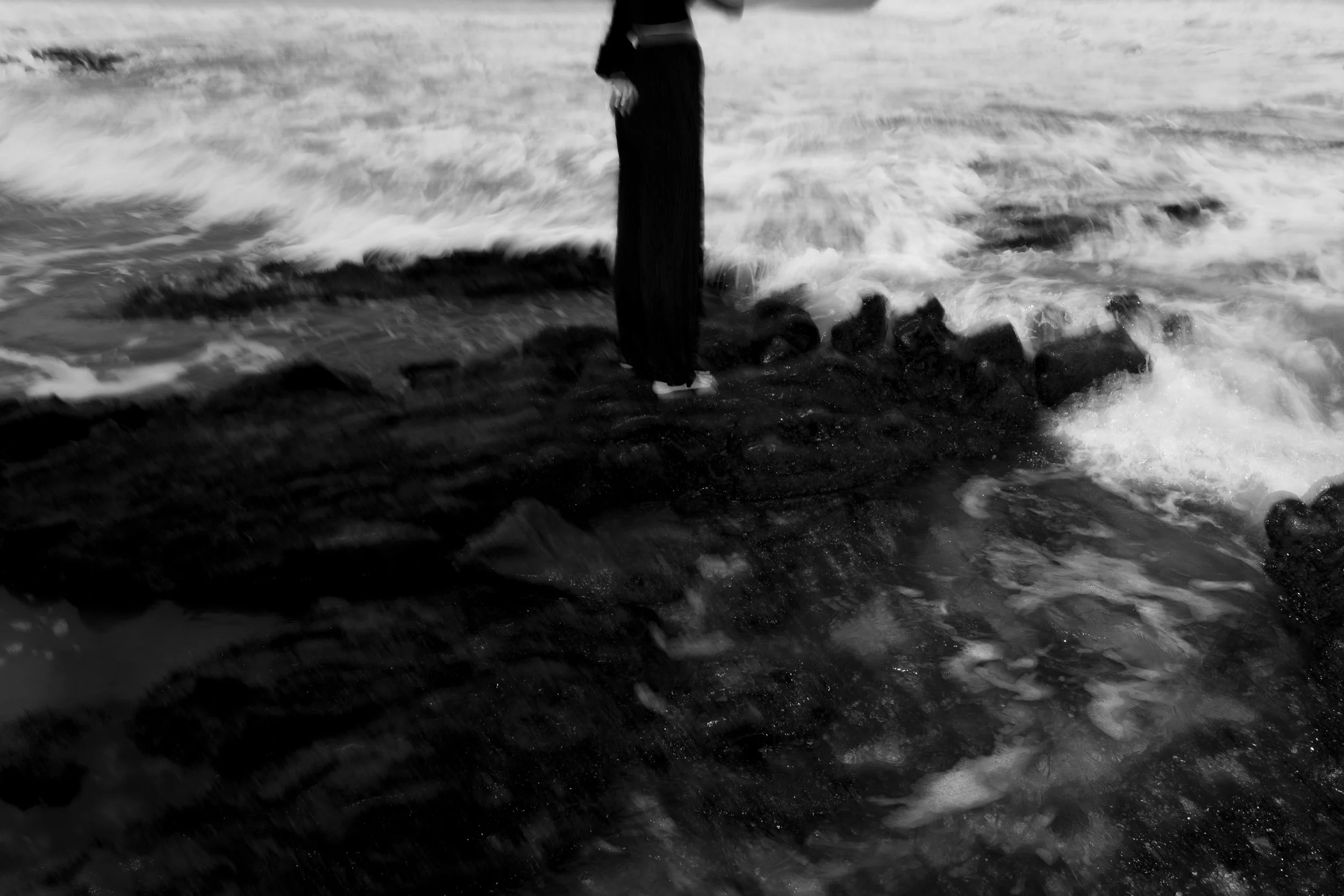 Black and white photo of a person standing on rocks near the ocean with waves crashing around them.