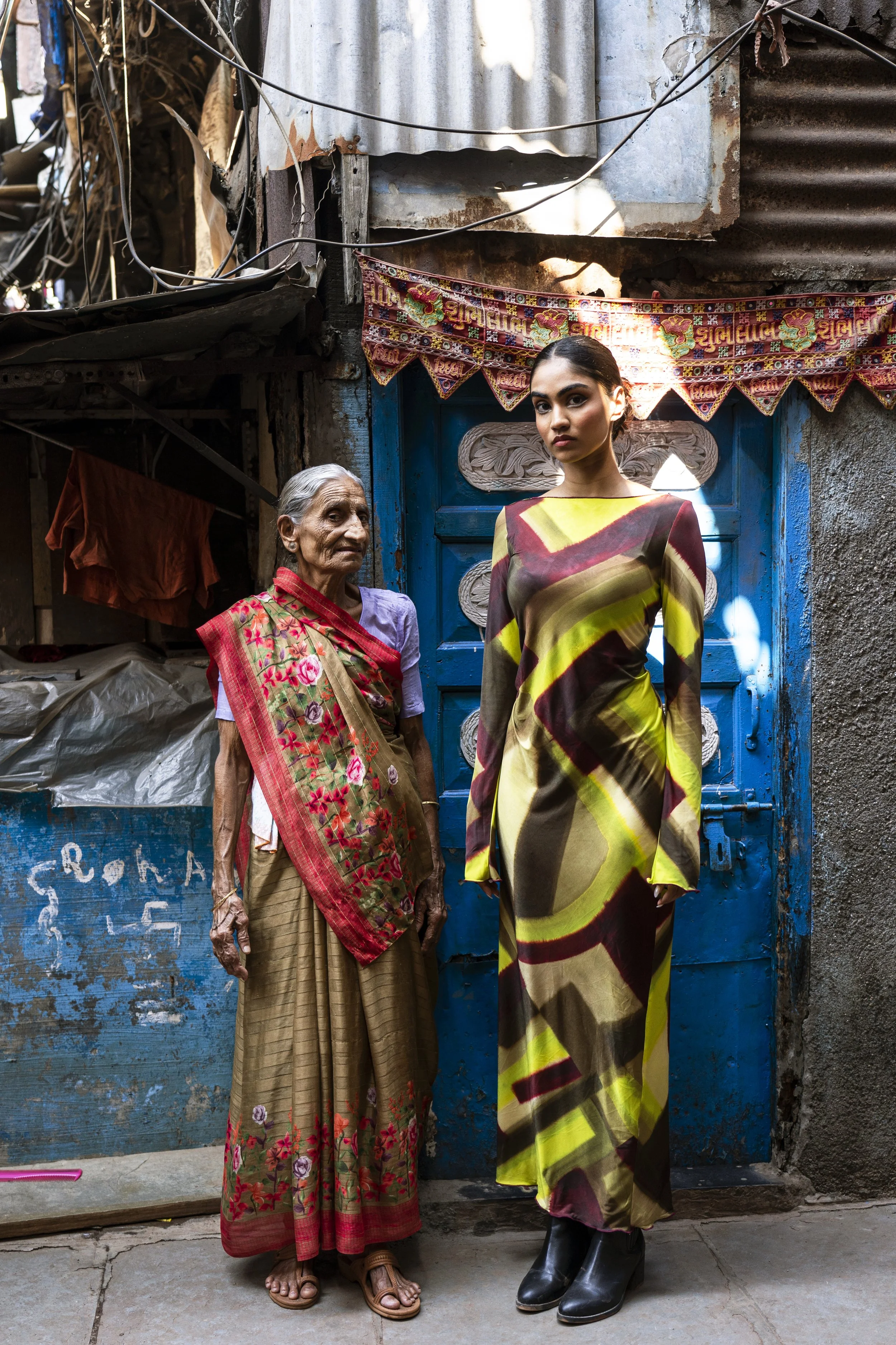 An elderly woman and a young woman standing in front of a blue door in an urban setting, with the older woman wearing traditional clothing with floral embroidery and the younger woman wearing a modern multicolored dress.