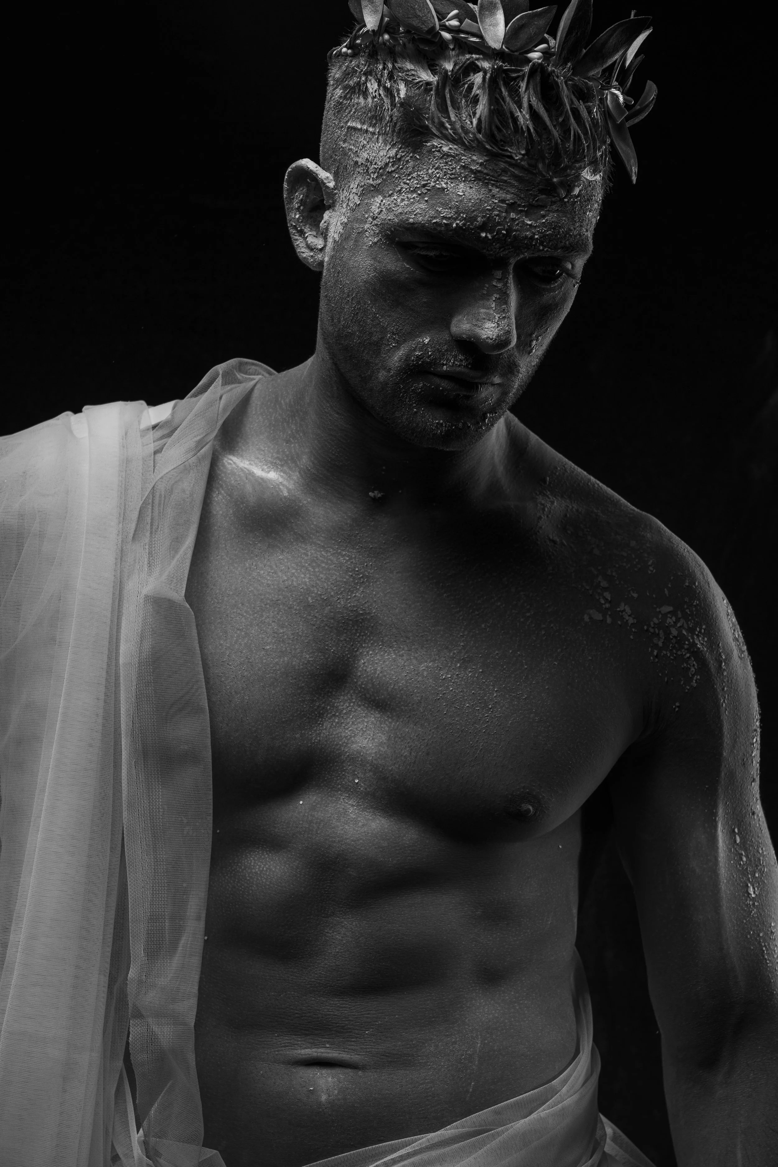 Black and white photograph of a male model with a serious expression, wet skin, and a crown of leaves on his head, partially draped in sheer fabric.