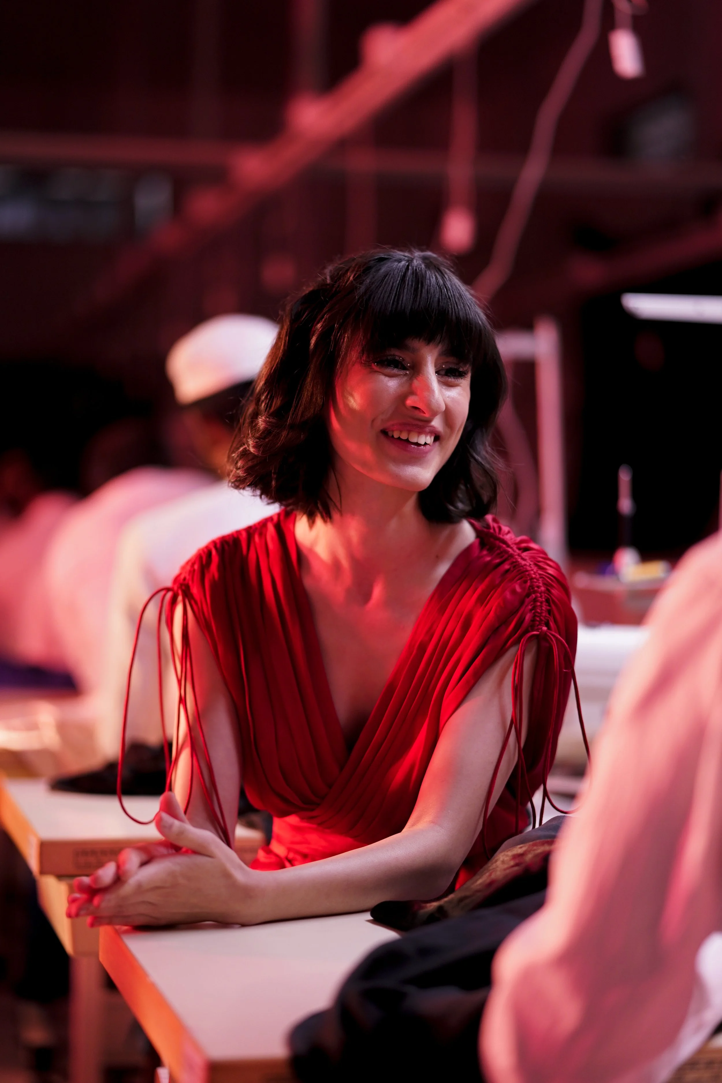 A woman with shoulder-length dark hair and a bright smile, wearing a red dress with pleats and drawstring details on the shoulders, sitting at a table in a dimly lit setting with red lighting.