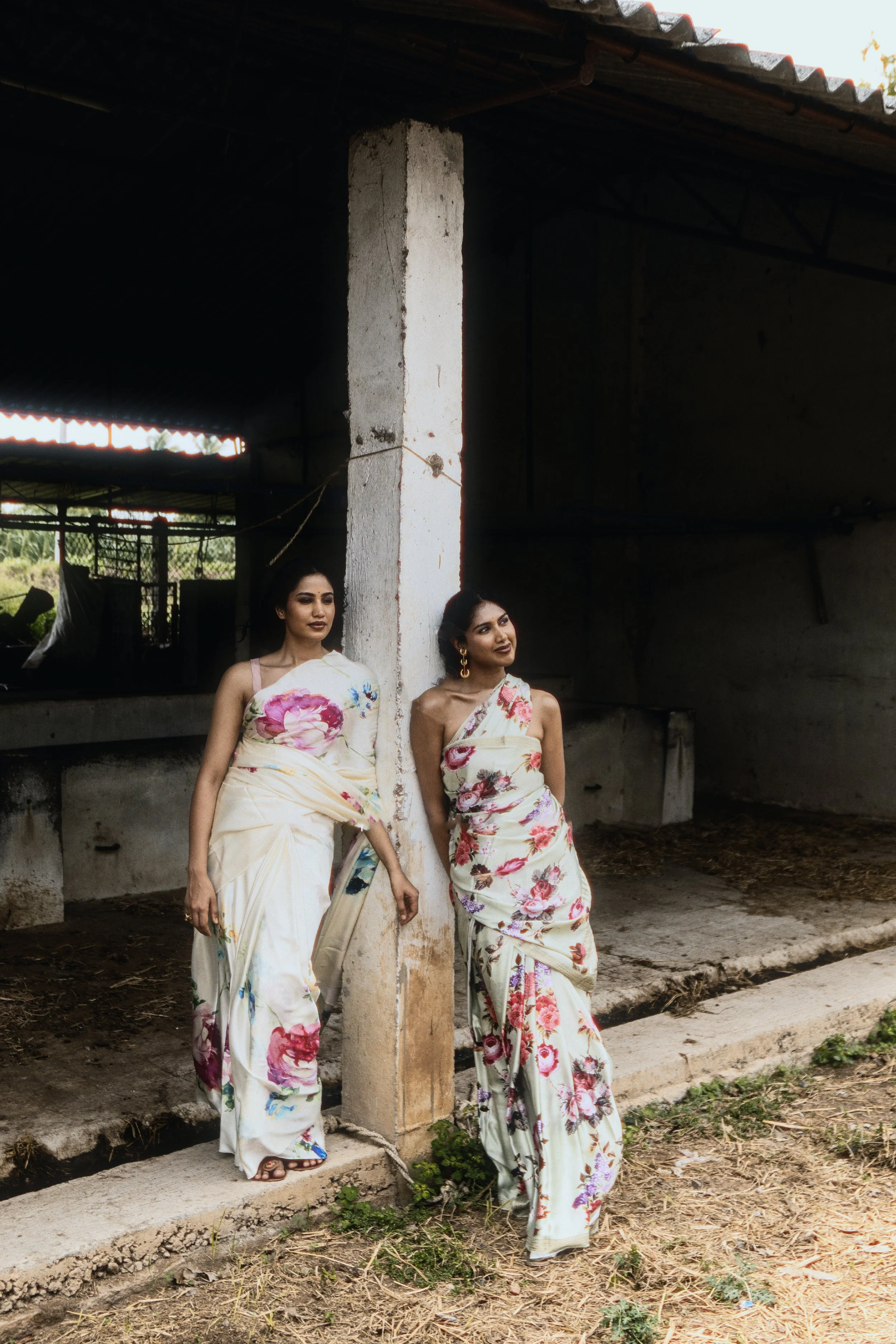 Two women in floral sarees standing outdoors, leaning against a concrete pillar, in front of a rustic building with a tiled roof.