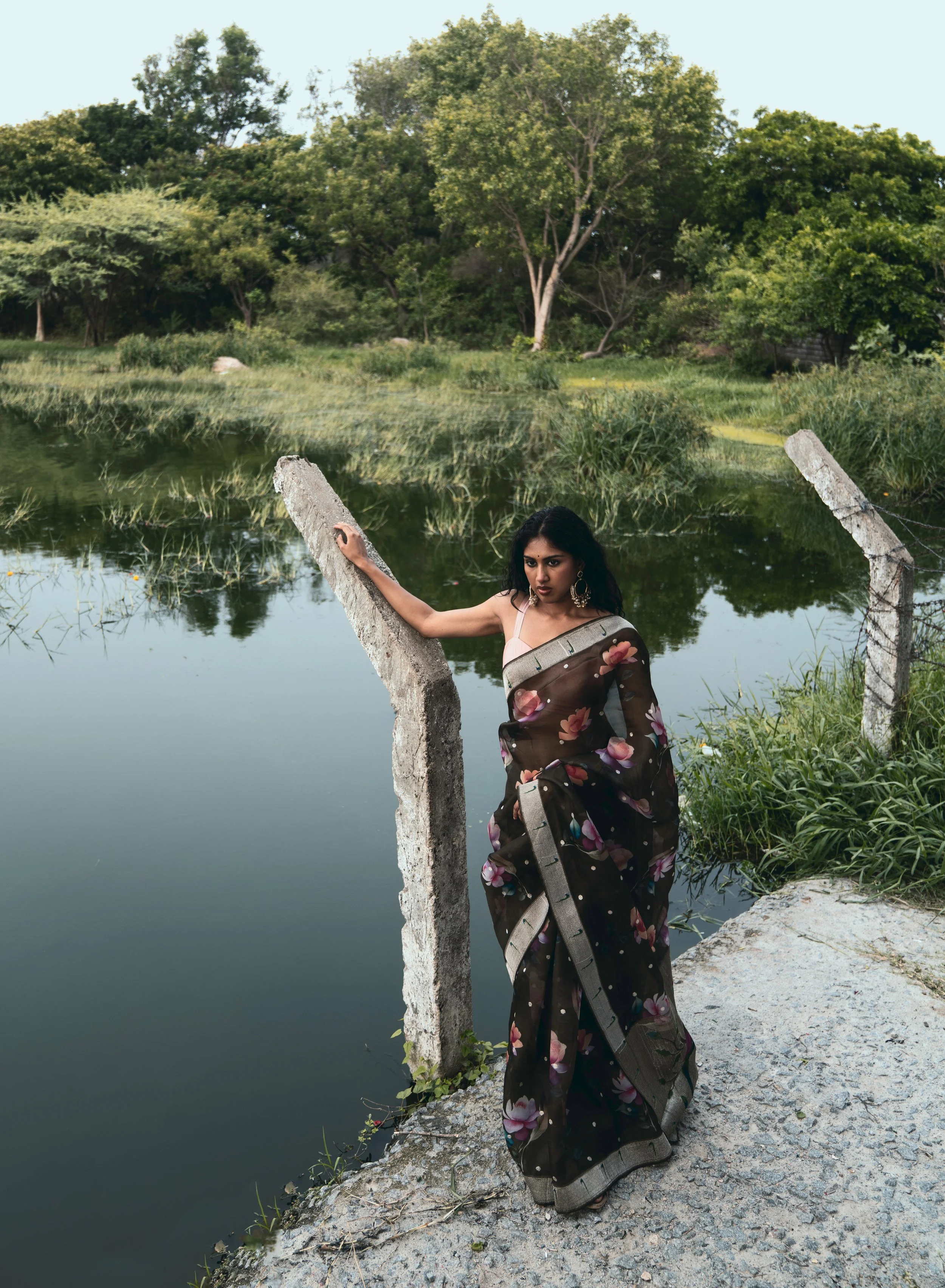 A woman in a floral saree standing on a stone path by a pond, holding a weathered fence post, with trees and grass in the background.