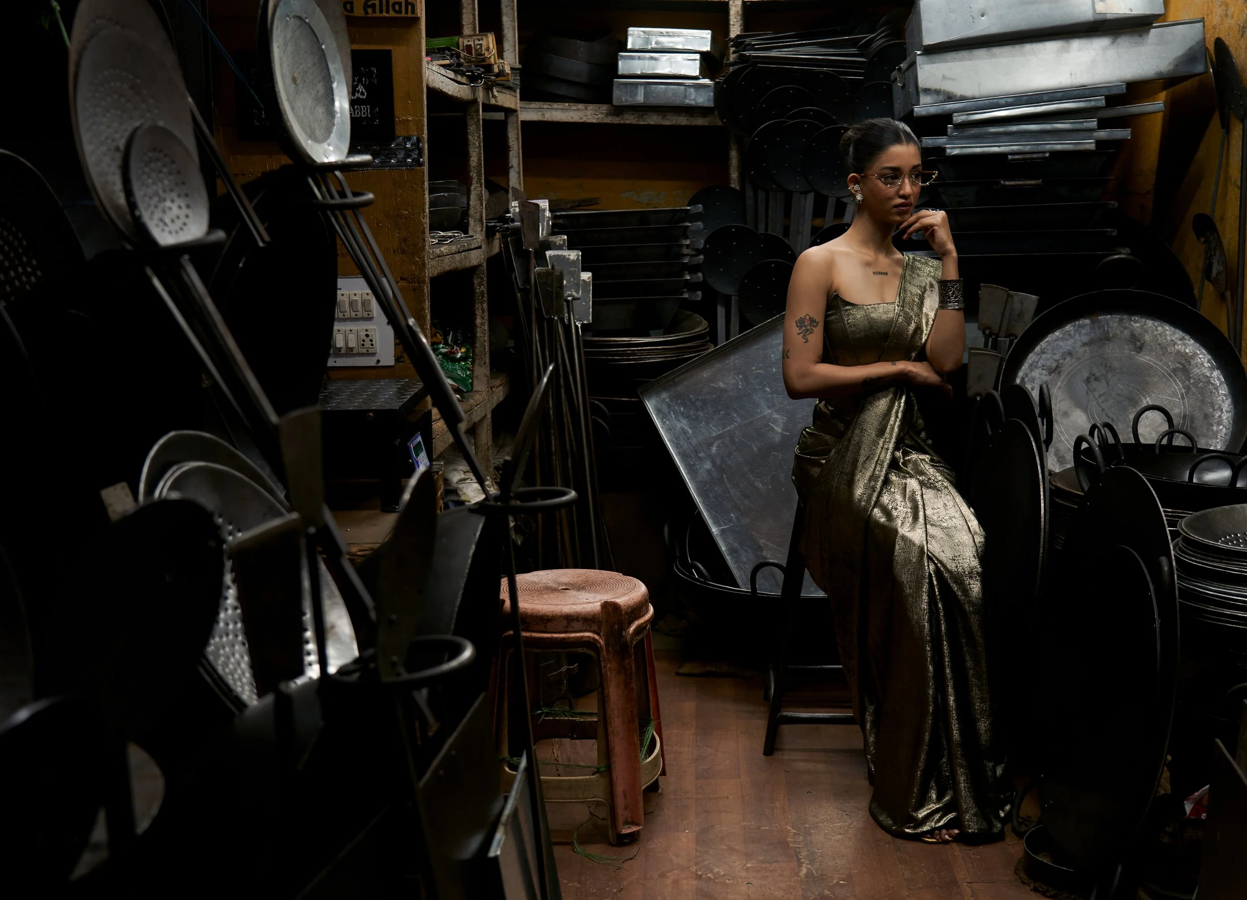 A woman in a gold sari sitting in a kitchen or storage room with metal kitchenware and utensils surrounding her.