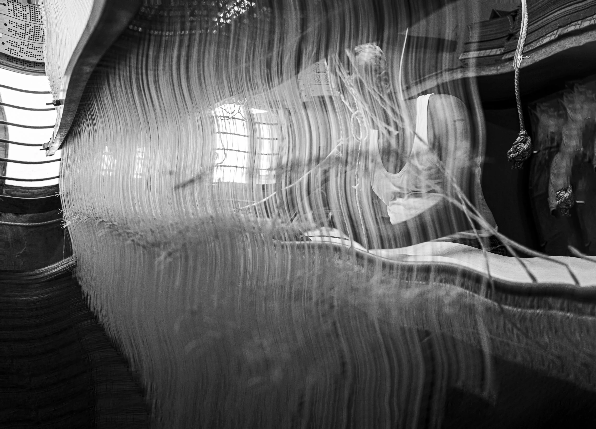 Black and white photo showing a cluttered room with a large, curved piece of furniture or wood, and a reflective surface creating abstract patterns. There is a person in the background wearing a sleeveless top, standing near a window with bars, with 