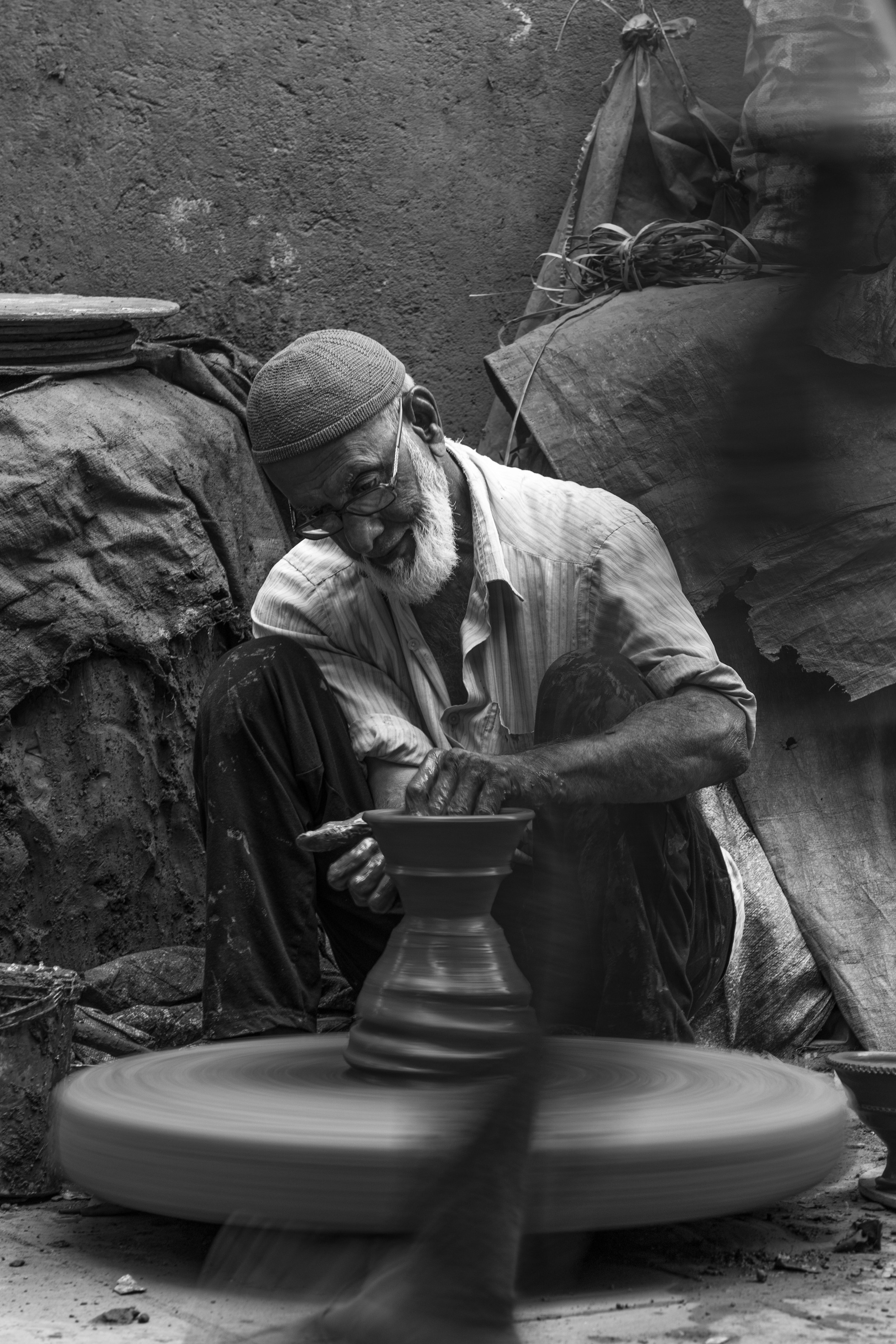 An elderly man wearing a cap and glasses, sitting cross-legged on a dirt floor, shaping pottery on a spinning wheel in a rustic workshop.