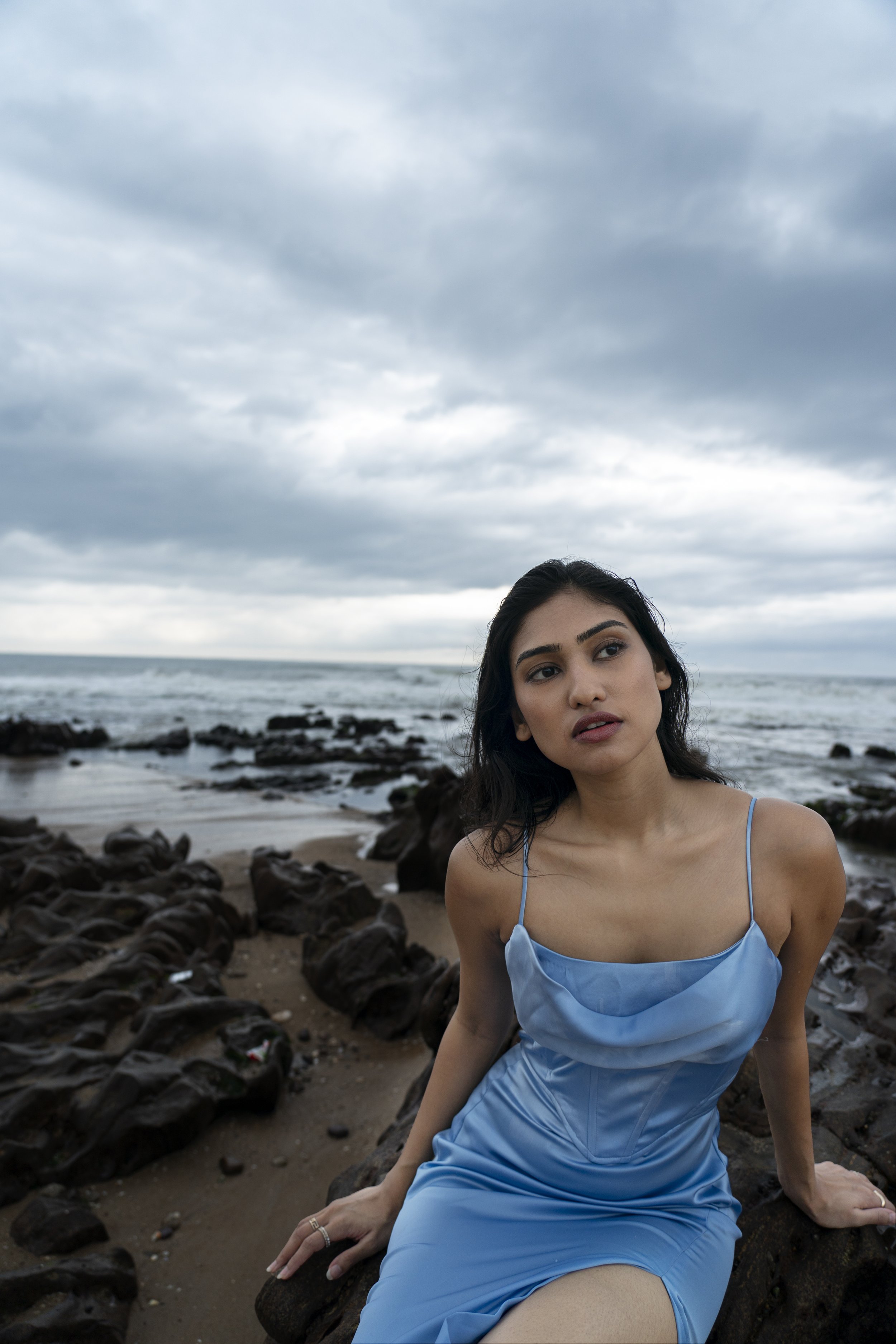 A woman in a blue dress sitting on rocks by the ocean under a cloudy sky.