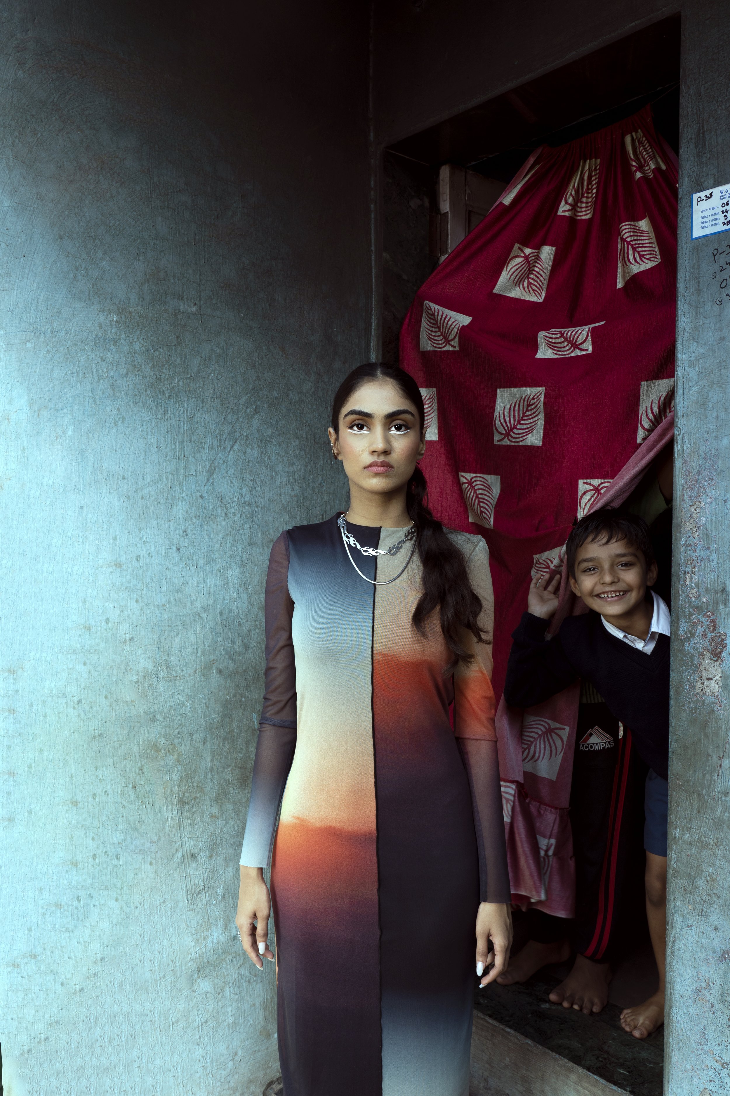 A young woman with long dark hair wearing a long multicolored dress, standing in front of a textured wall. Behind her, a boy is peeking out from a doorway decorated with a red curtain with white leaf patterns, smiling and waving.