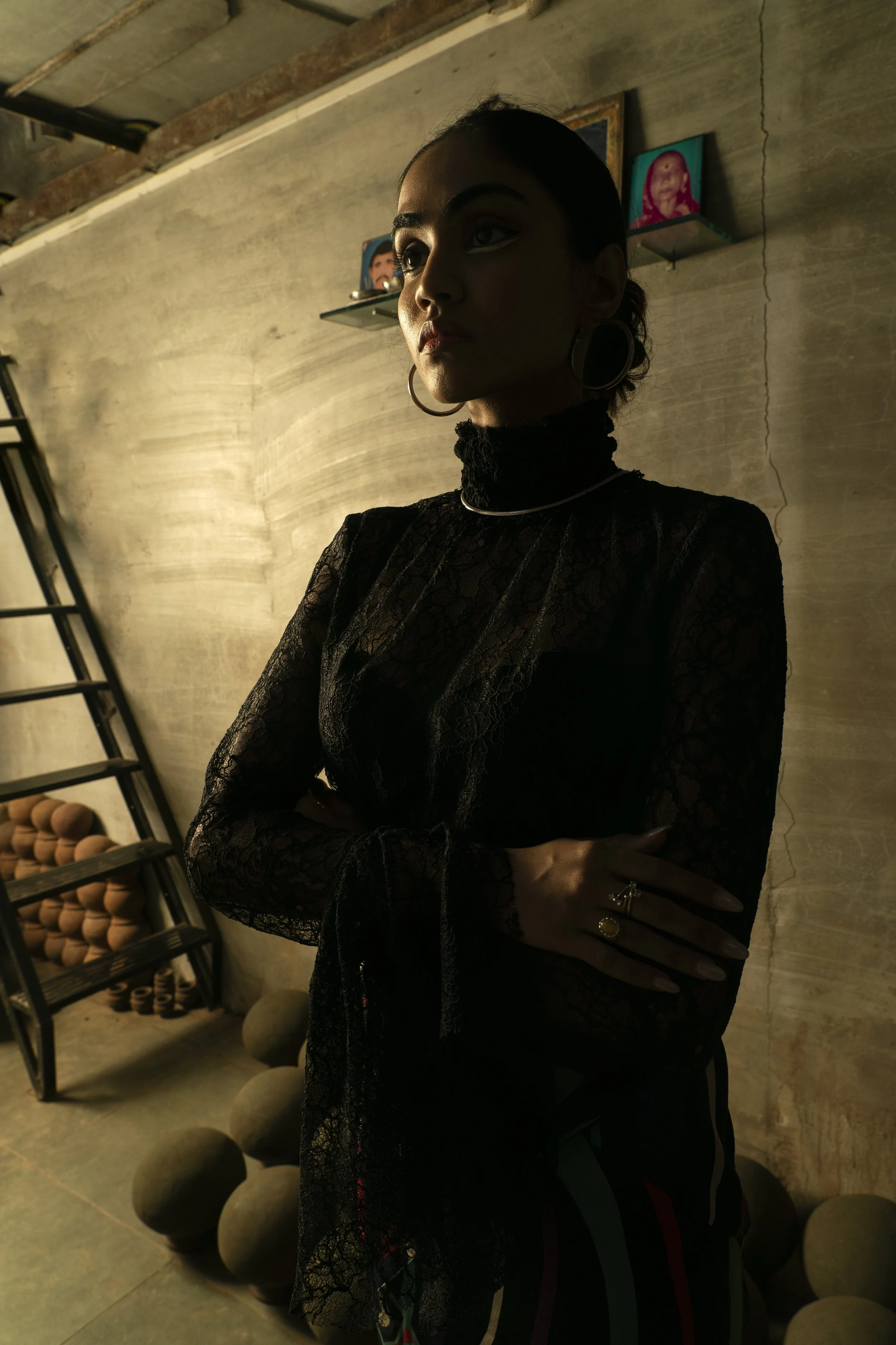 A woman with crossed arms wearing a black lace top, earrings, and rings standing in a room with a textured wall, a ladder, and shelves with pottery in the background.