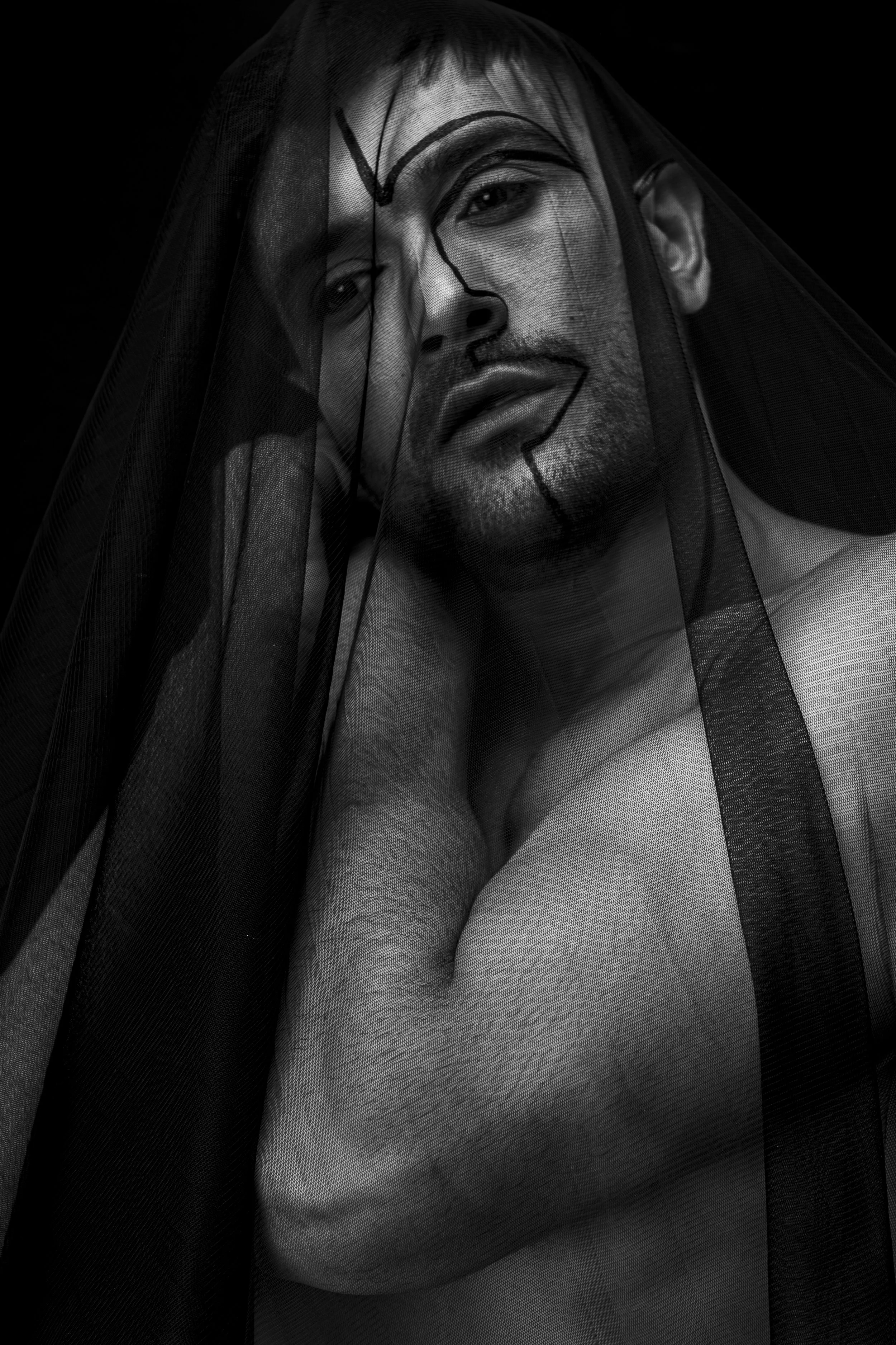 A Black and white portrait of a young man with a veil over his face, which is painted with a black crown and other markings.