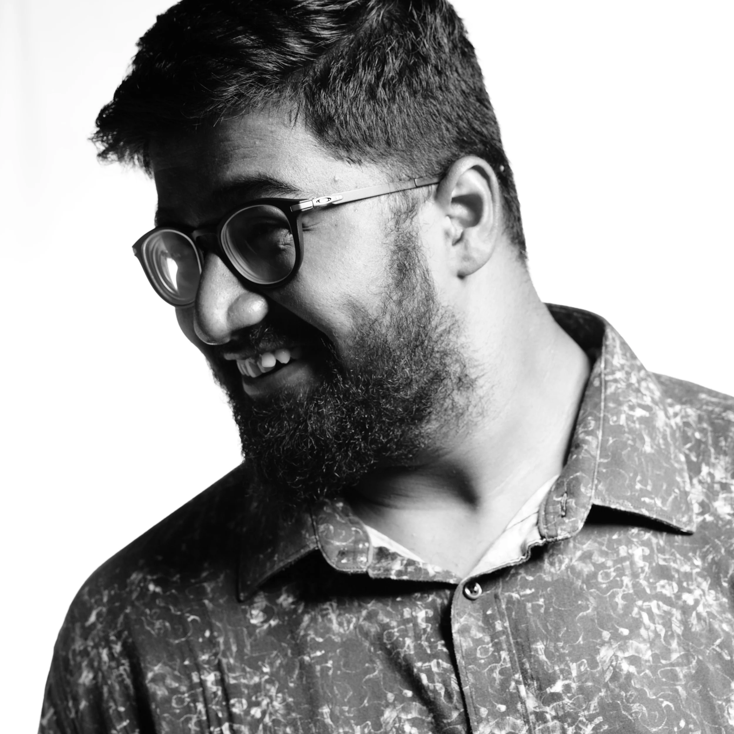 Black and white portrait of a smiling man with glasses, a beard, and short hair, wearing a patterned button-up shirt.