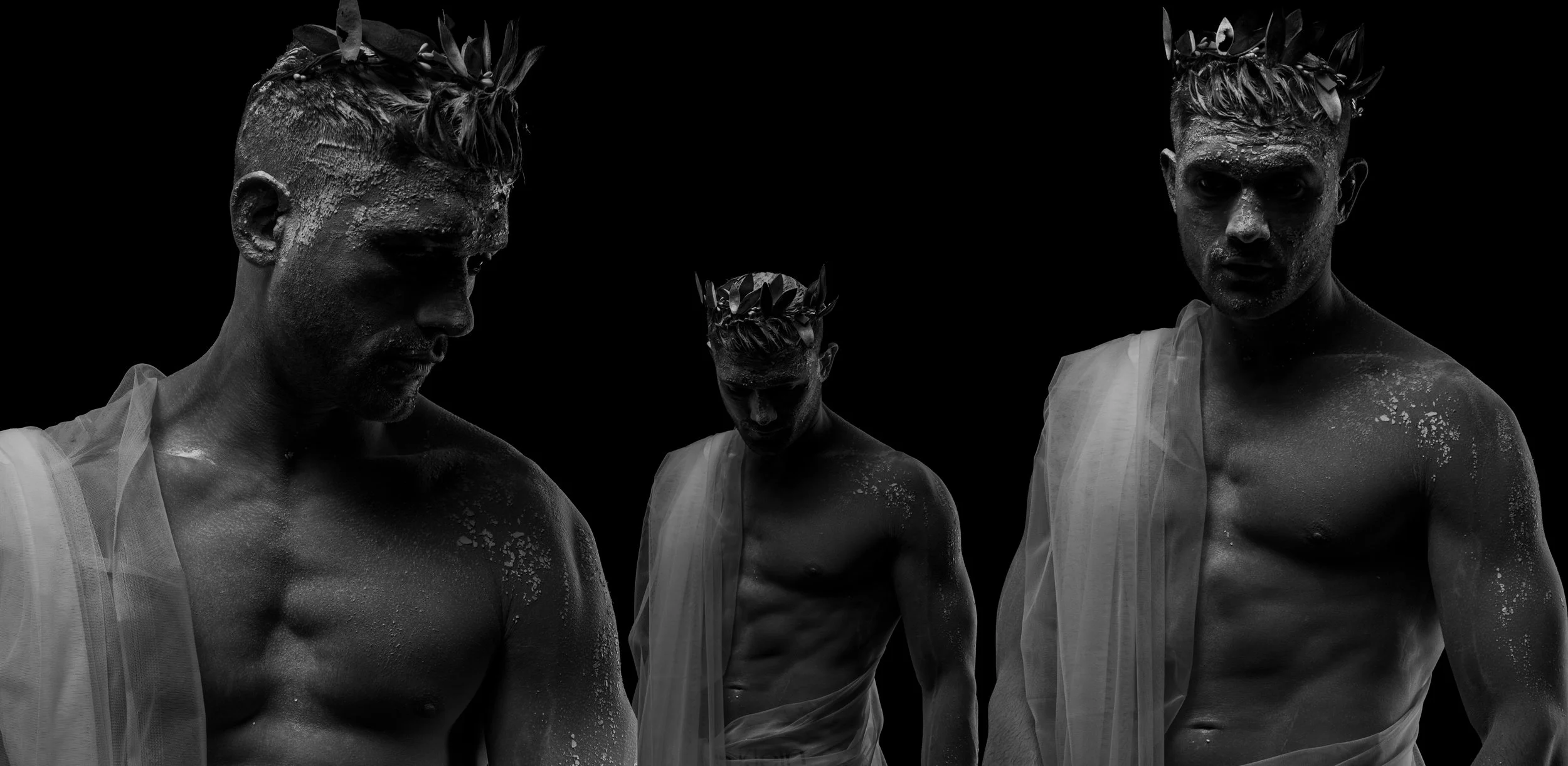 Black and white photo of three shirtless men with short hair, wearing laurel wreaths on their heads and draped in light, sheer fabric, standing against a dark background.